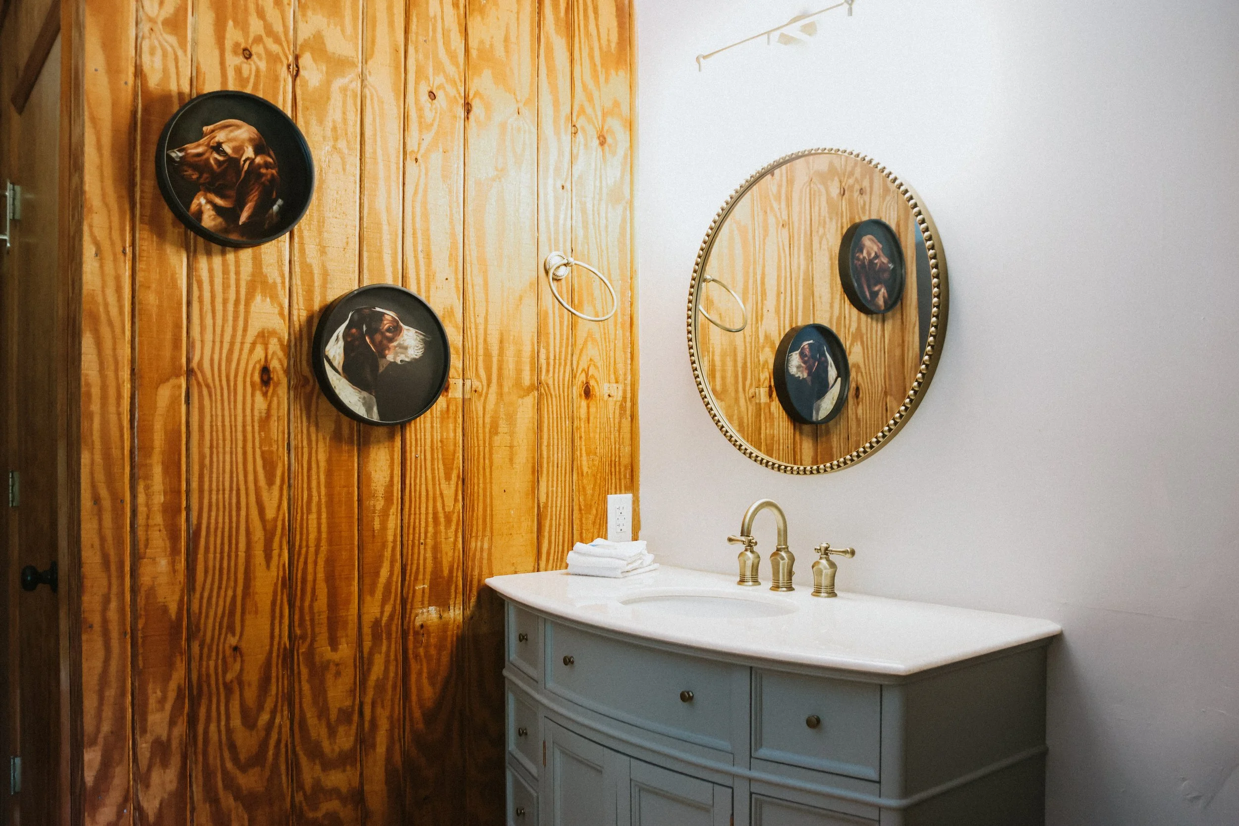 Bathroom with a wooden-paneled wall, oval mirror, and framed portraits of dogs on the wall.