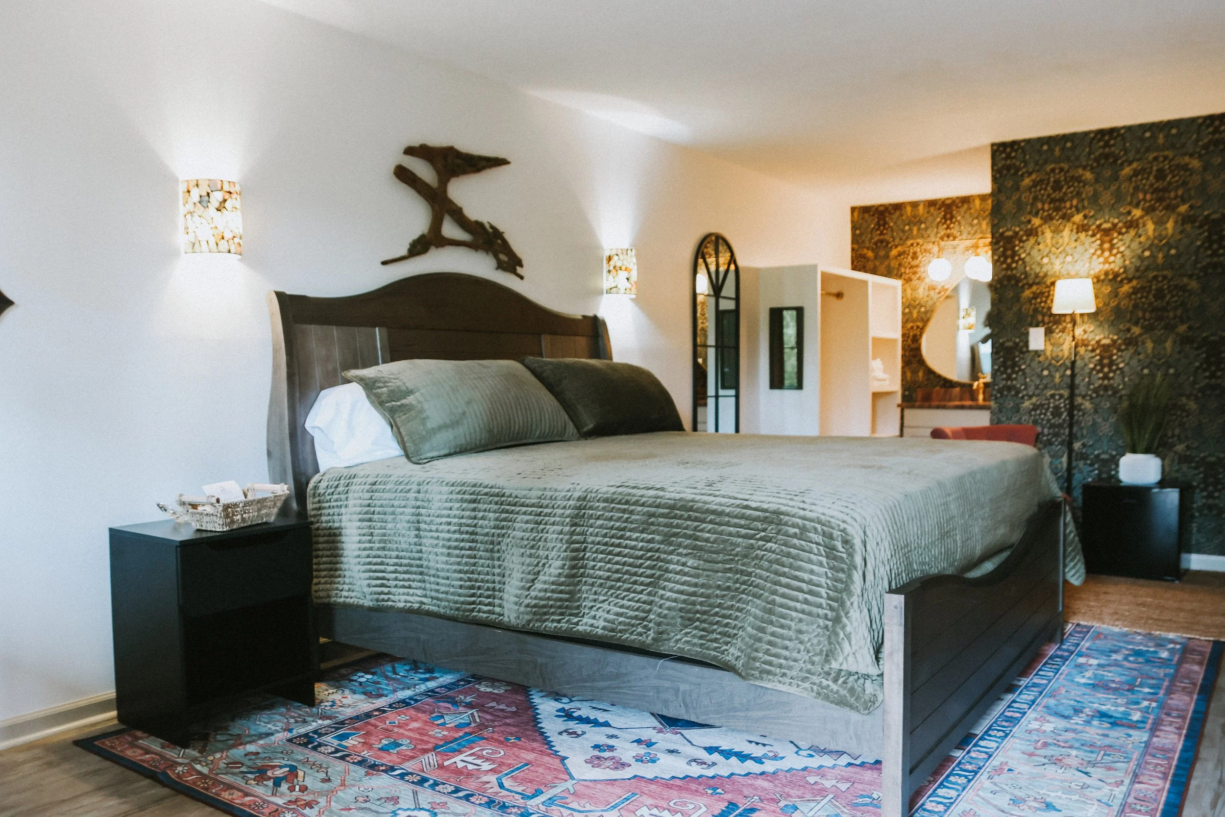A bedroom with a large bed covered in a beige quilt, with multiple pillows, a dark wooden headboard, and a colorful oriental rug underneath. There are wall-mounted lamps, a black side table with a basket, and a decorative branch above the bed. The ro
