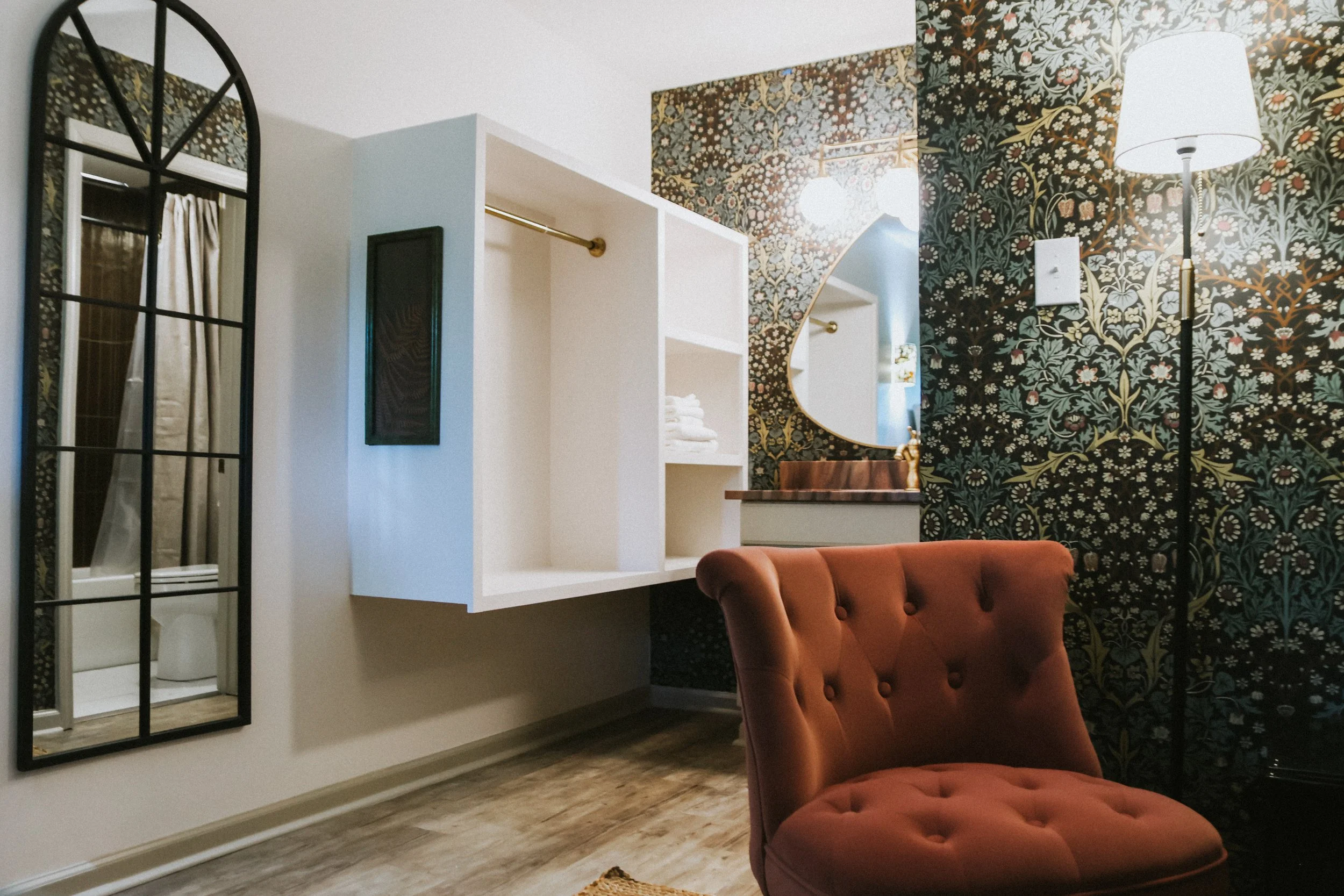Interior of a room with floral wallpaper, a tall floor lamp, a large mirror, a white open closet with towels, a vintage-style chair, and a mirror on the wall showing a part of a bathroom.
