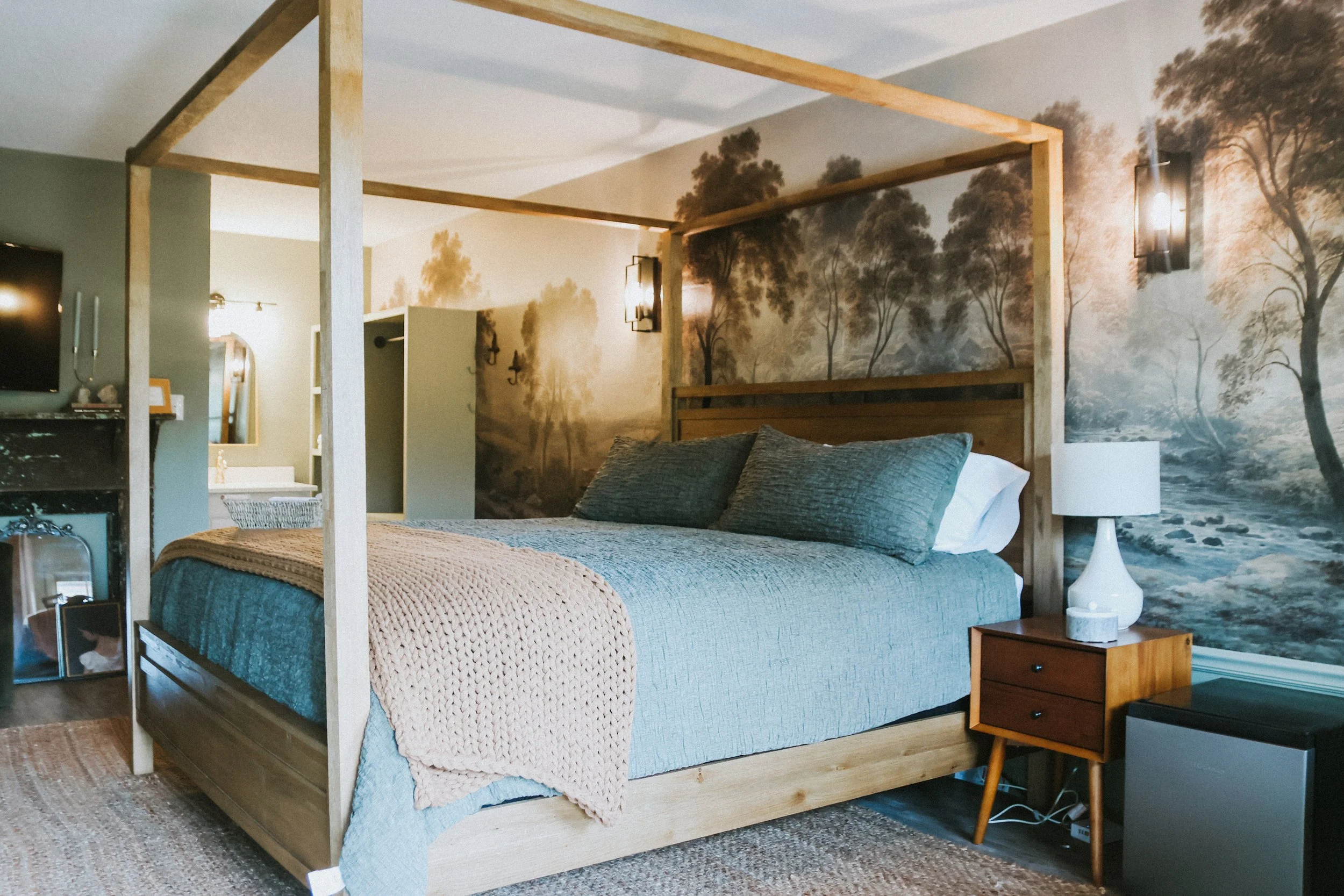 A bedroom with a wooden four-poster bed, blue bedding, gray pillows, and a beige knit throw blanket. The room has a nature scene mural with trees and a river on the wall. There is a nightstand with a white lamp, a small black appliance, and a beige r