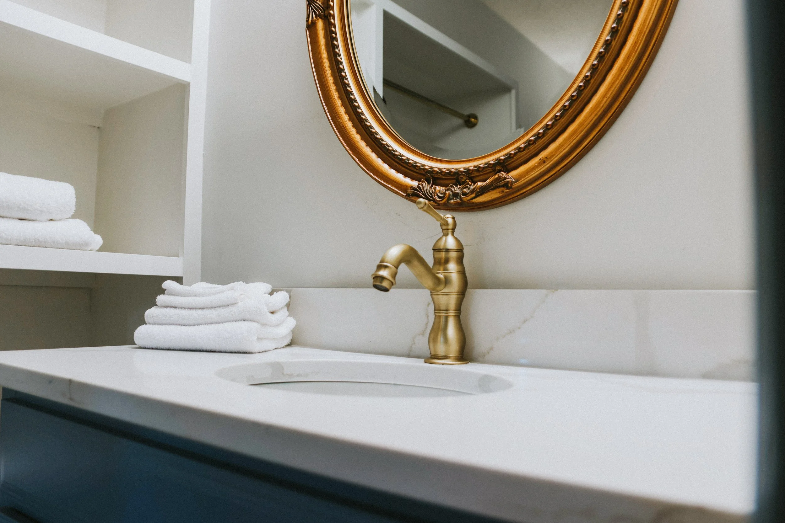 A bathroom sink with a gold faucet, a round decorative mirror above, and white folded towels on a shelf to the left.