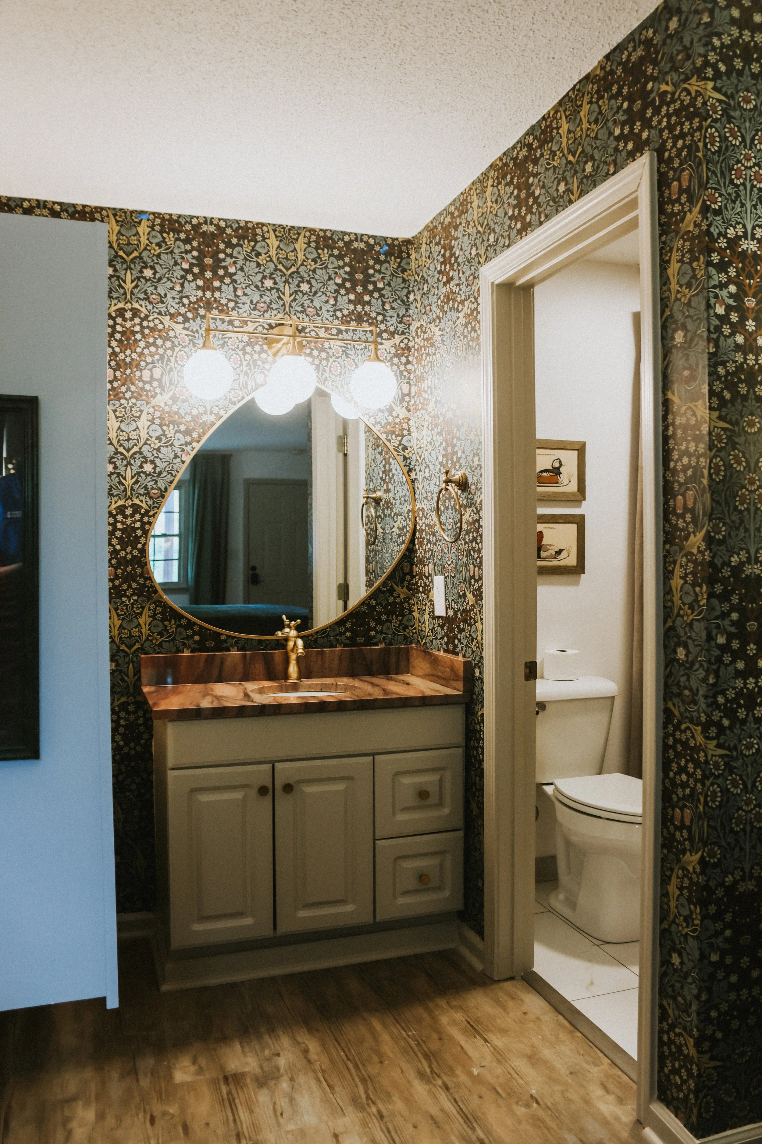 Bathroom with a vanity mirror, sink, and marble countertop, next to a toilet, with floral wallpaper and wooden floor.
