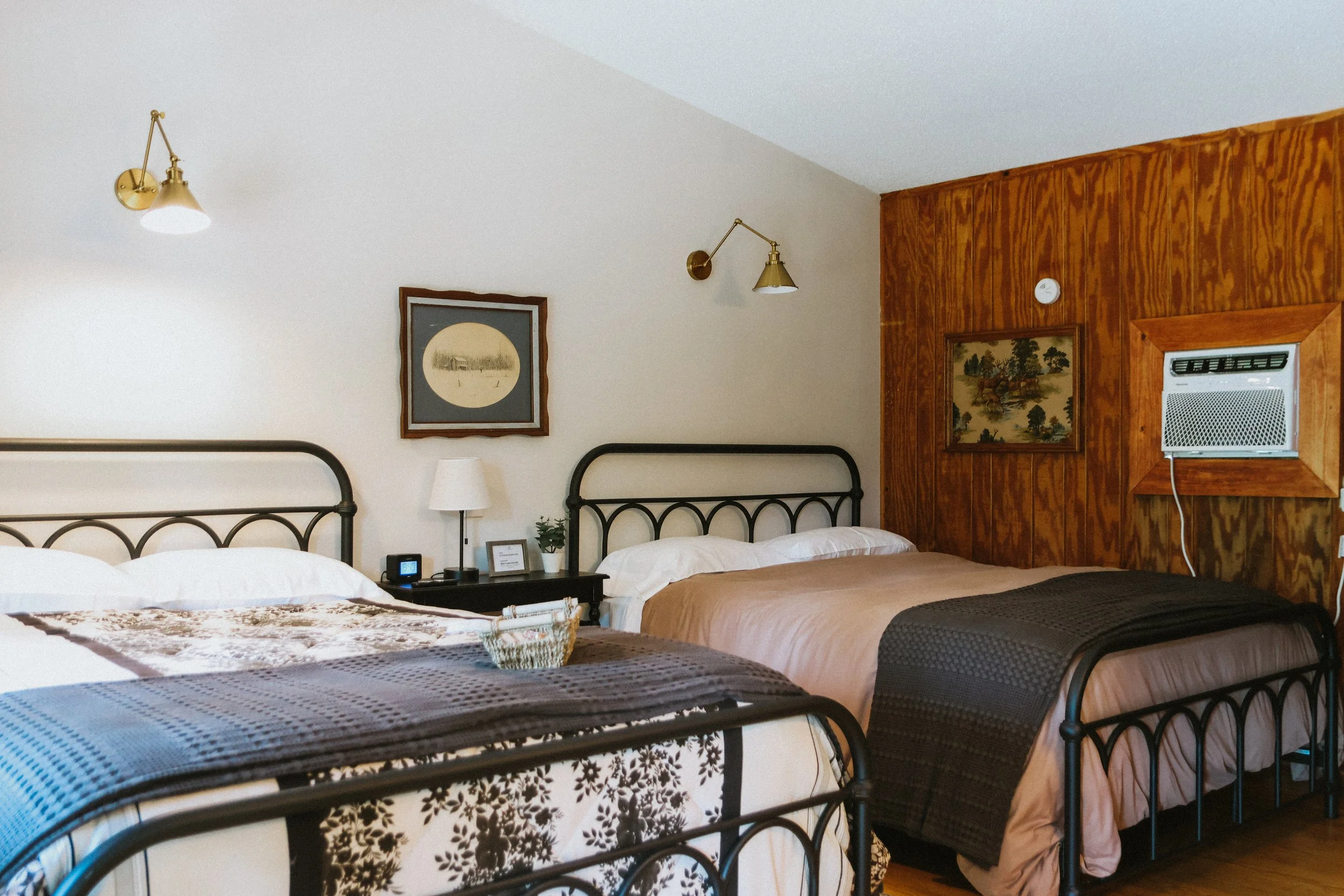 A bedroom with two beds featuring black metal frames, brown and beige bedding, and decorative black and white blankets. A black nightstand between the beds has a white lamp, a digital clock, and small potted plants. The walls are half white and half 