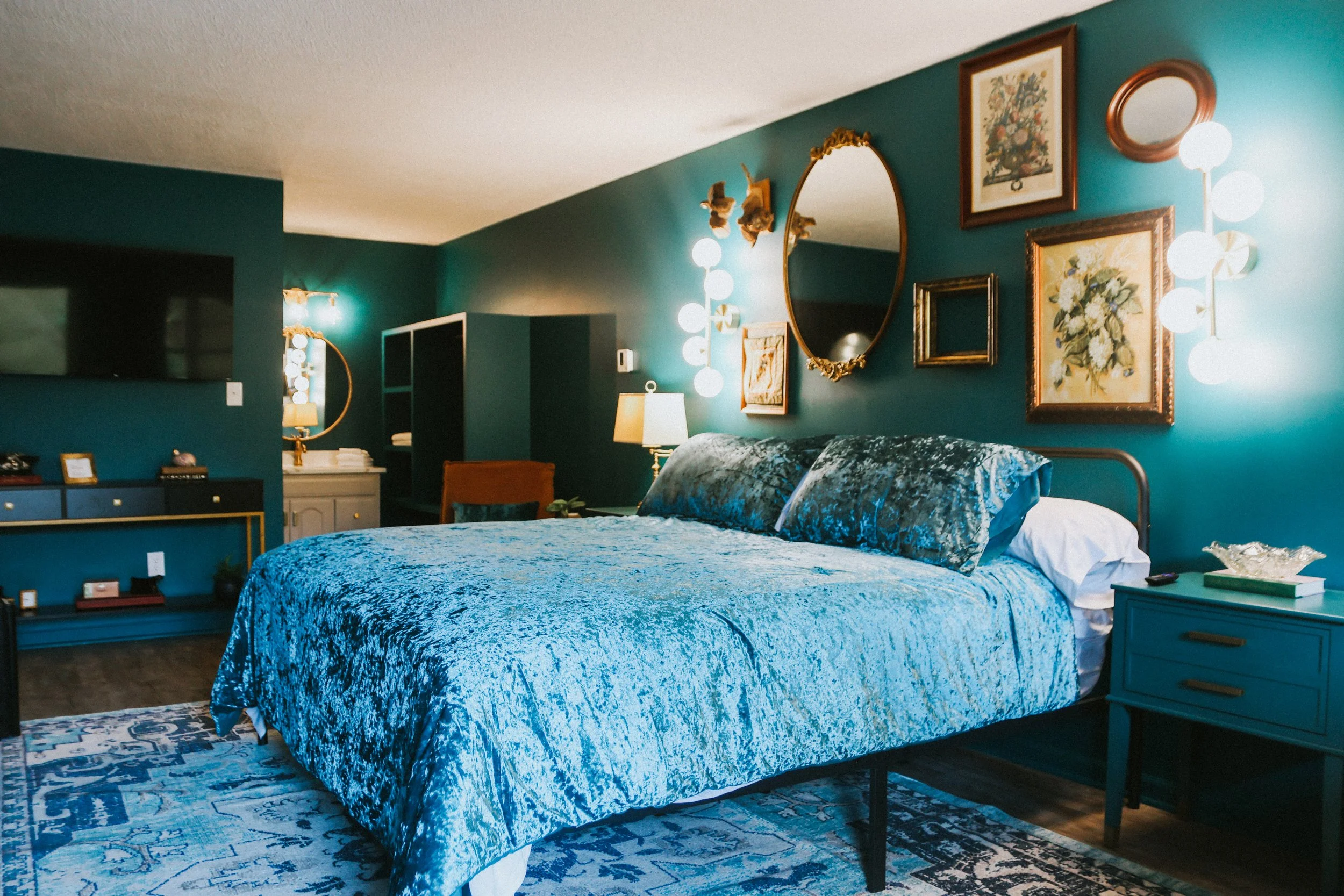 A bedroom with teal walls, a bed with a blue crushed velvet bedspread, and matching pillows. There is a blue nightstand with a dish and books, and framed artwork on the wall. The room has a mirror, lamps, and wall sconces with globed lights overhead.