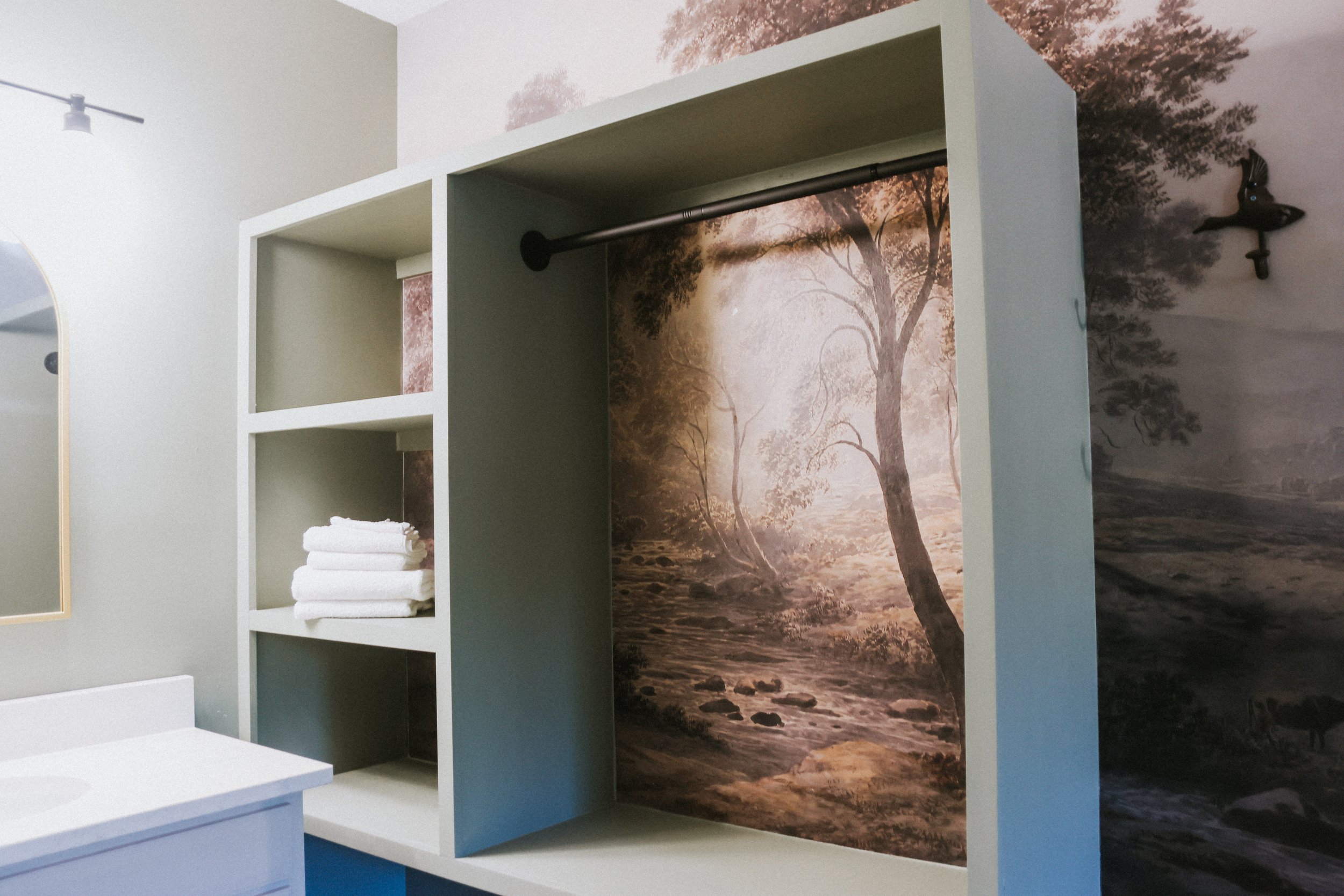 Part of a bathroom with a mural of trees and a river on the wall, a white shelf with towels, and a bathroom sink.