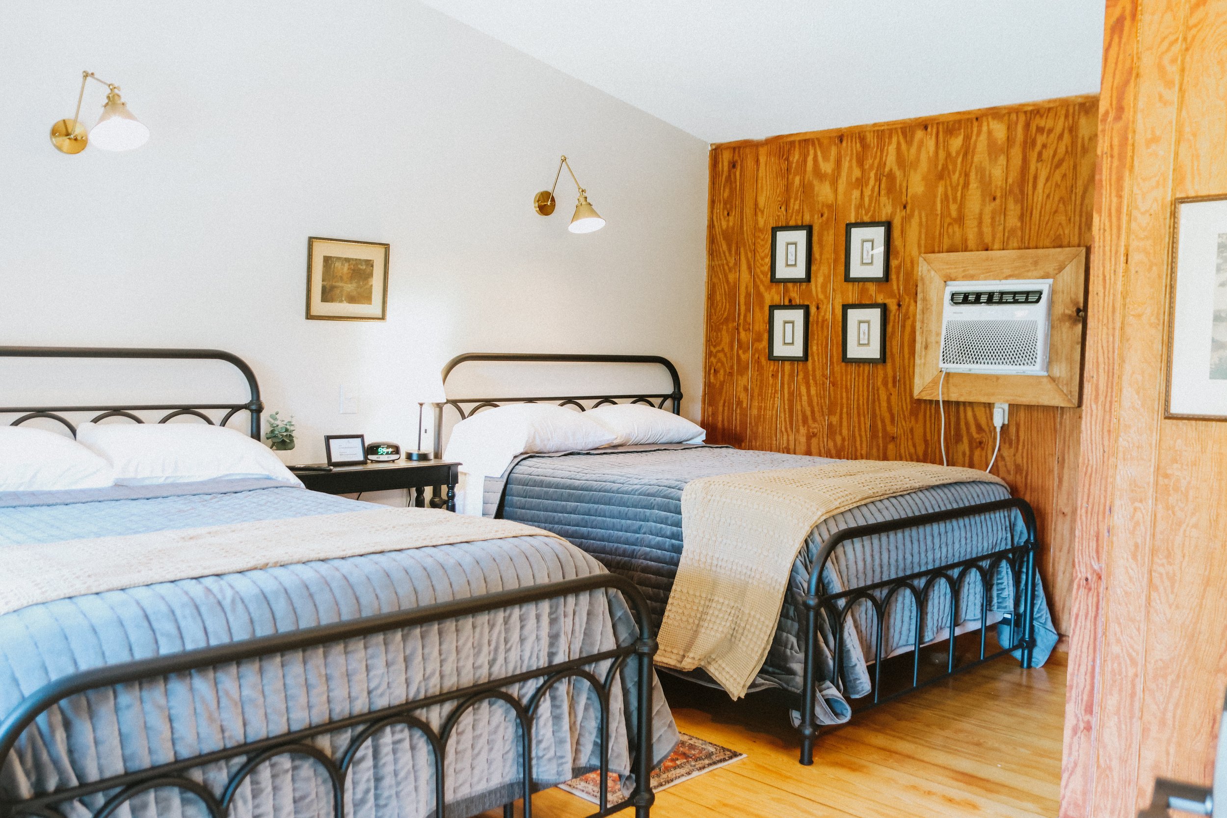 Two beds with metal frames, quilted bedspreads, pillows, and blankets in a bedroom with wooden paneling on one wall and white walls; wall-mounted air conditioner, framed pictures, and bedside table with clock and lamp.