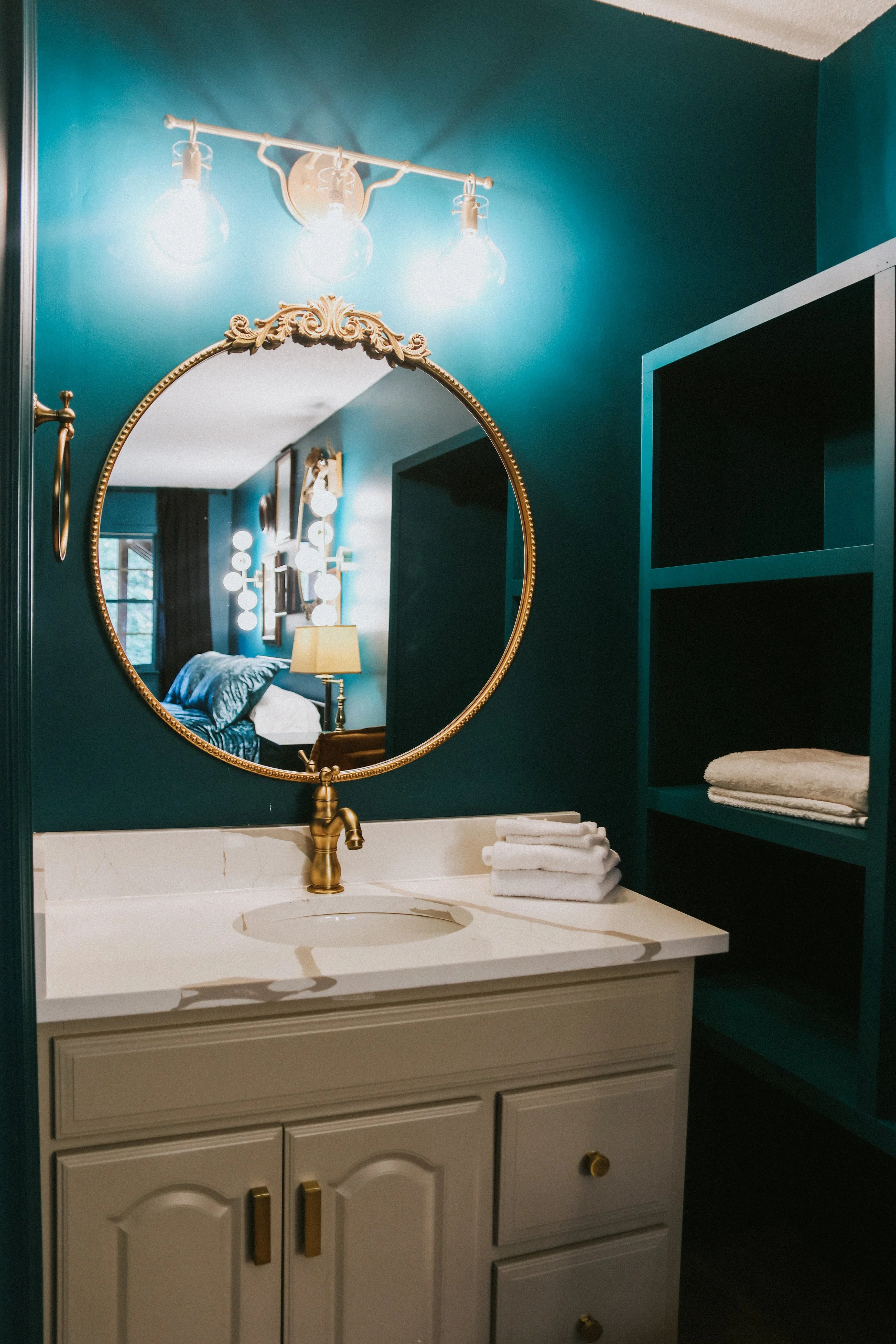 Bathroom vanity with a white cabinet, brass faucet, oval mirror, blue walls, and shelves with folded towels.