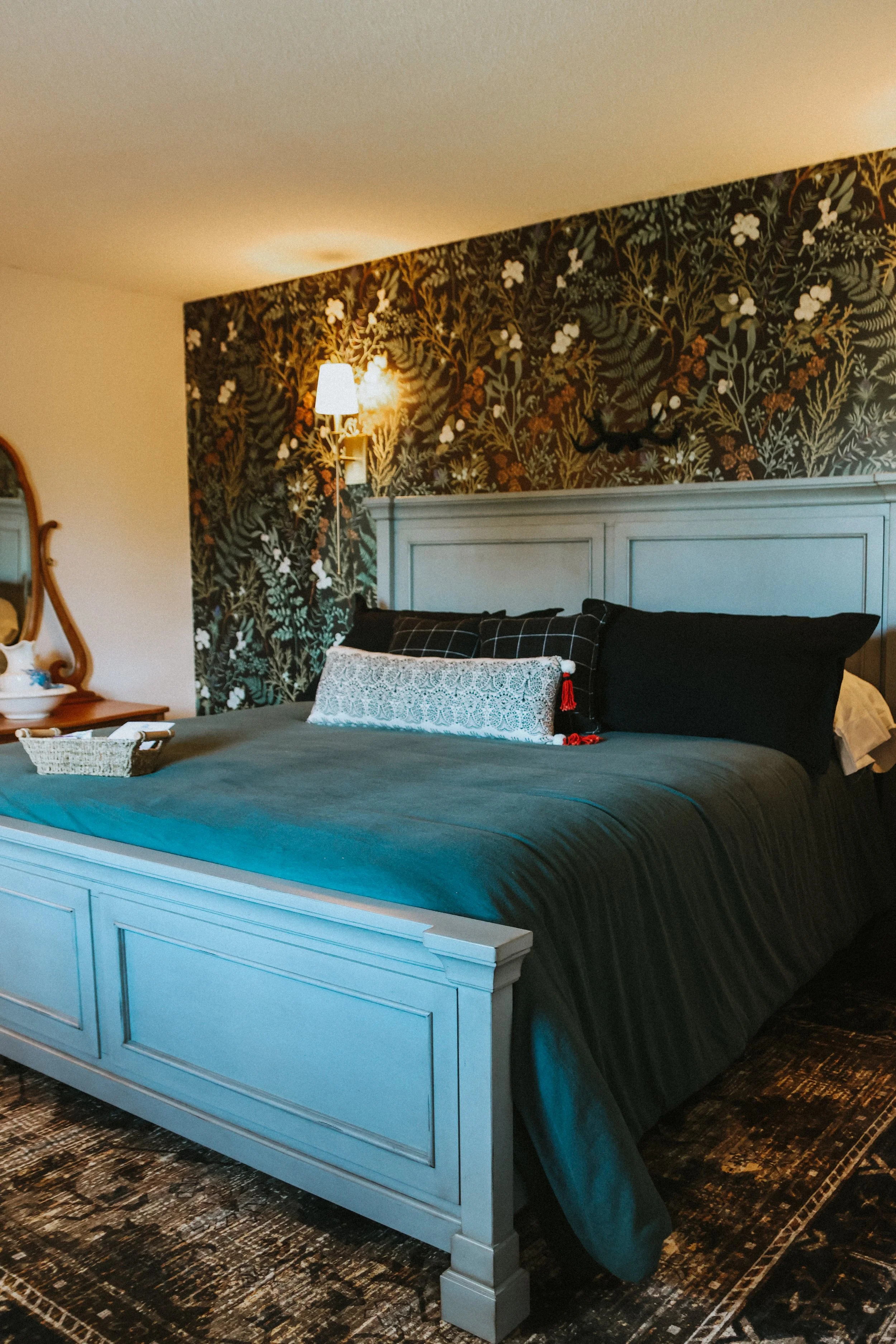 A neatly made bed with dark bedding, decorative pillows, and a tufted headboard in a bedroom with floral wallpaper and soft lighting.