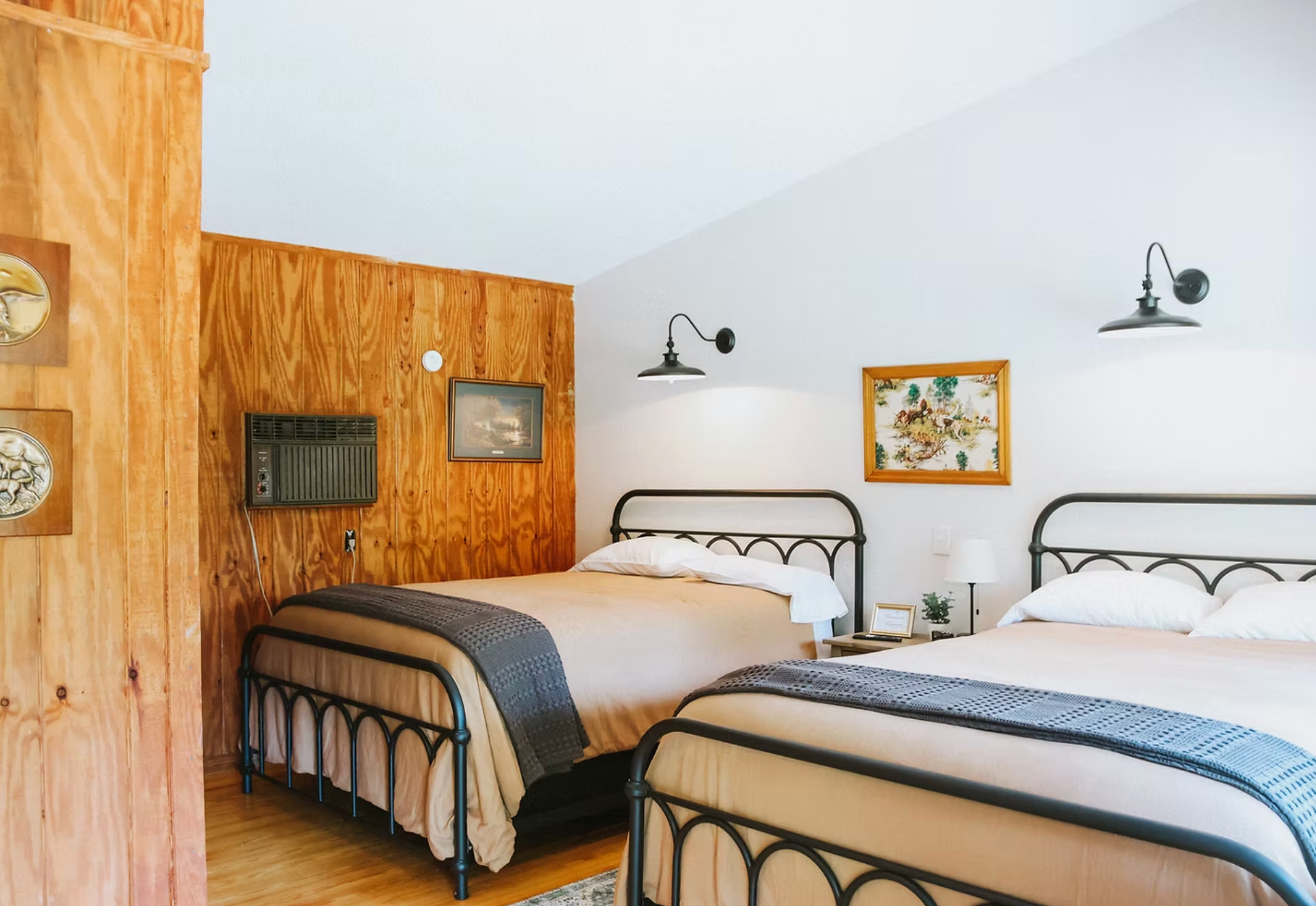 A cozy bedroom with two metal frame beds, a wooden-paneled wall, framed artwork, wall-mounted lights, a nightstand with a small lamp and plant, and hardwood floor.