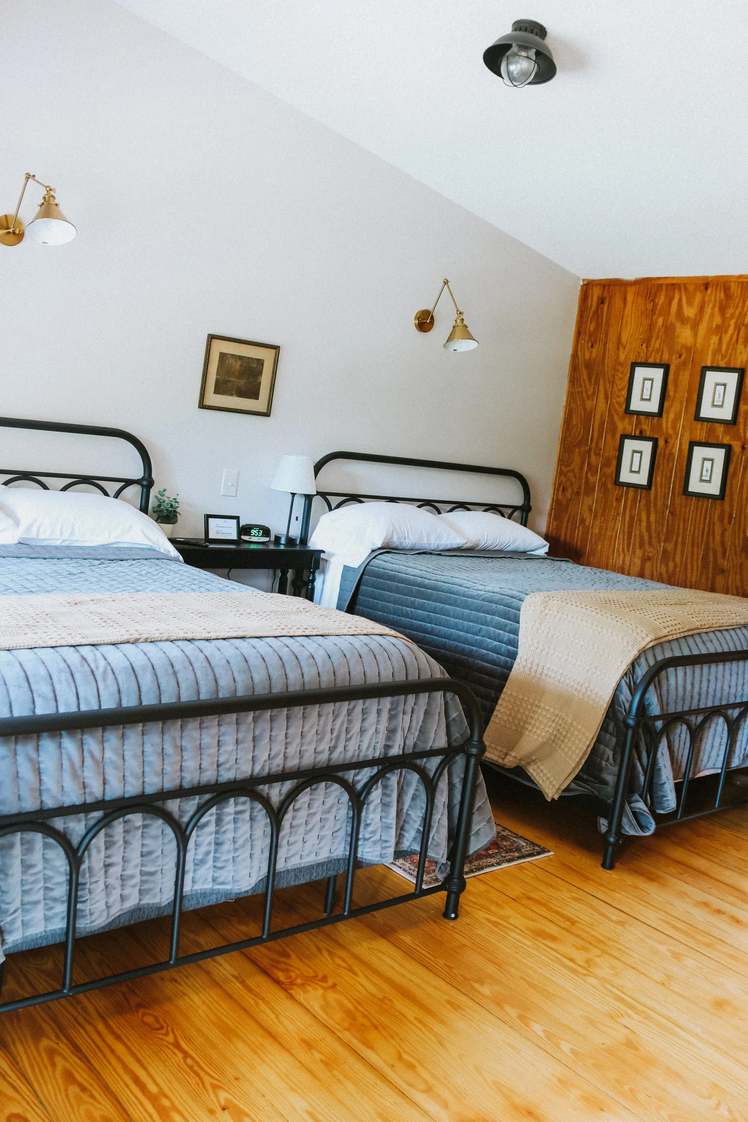 Two beds with metal frames and striped bedding in a cozy bedroom, with a wooden wall, wall-mounted lights, and framed pictures on the wall.