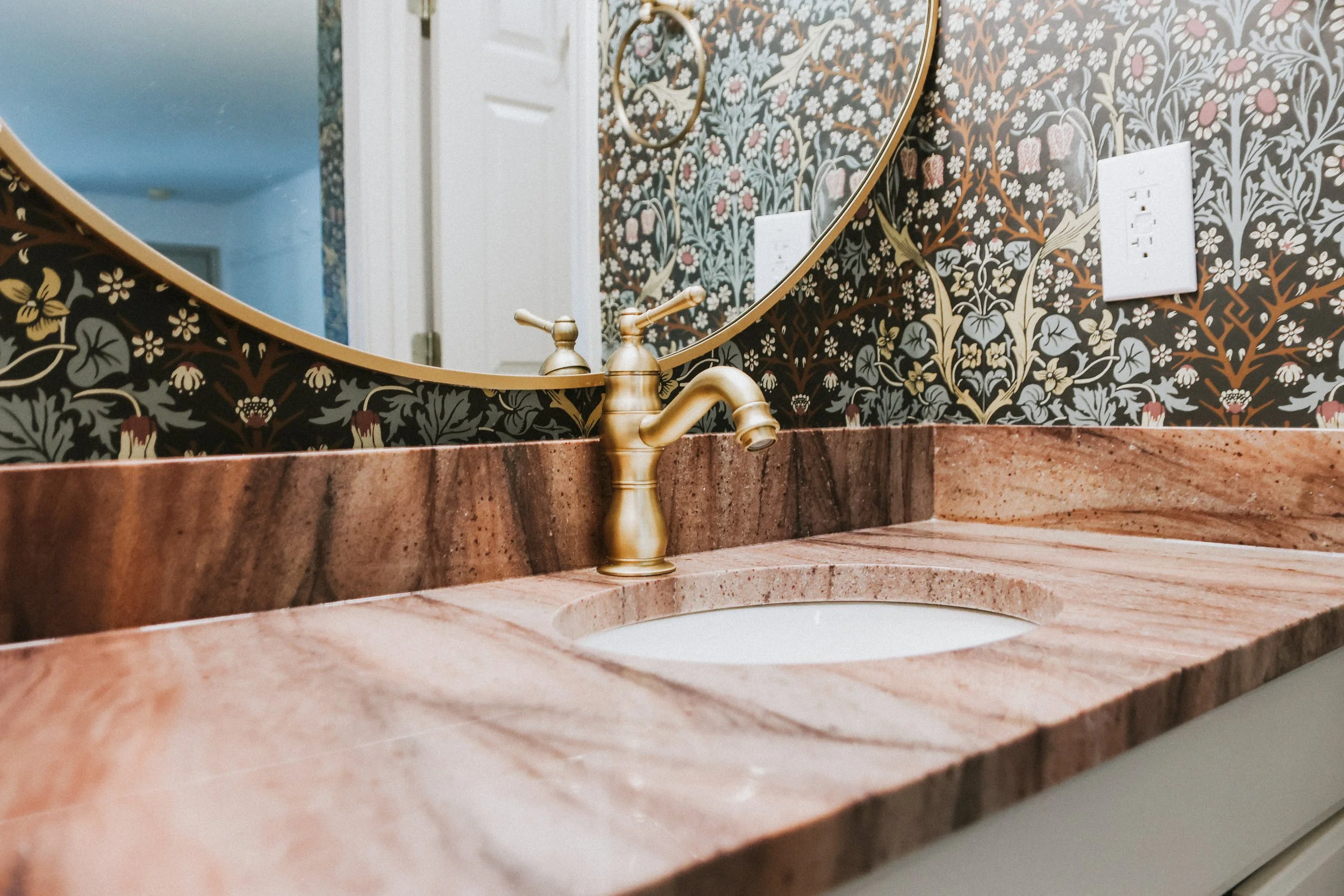 Close-up of a bathroom sink with a pinkish marble countertop, gold faucet, and a decorative floral wallpaper with an oval mirror reflected in the background.