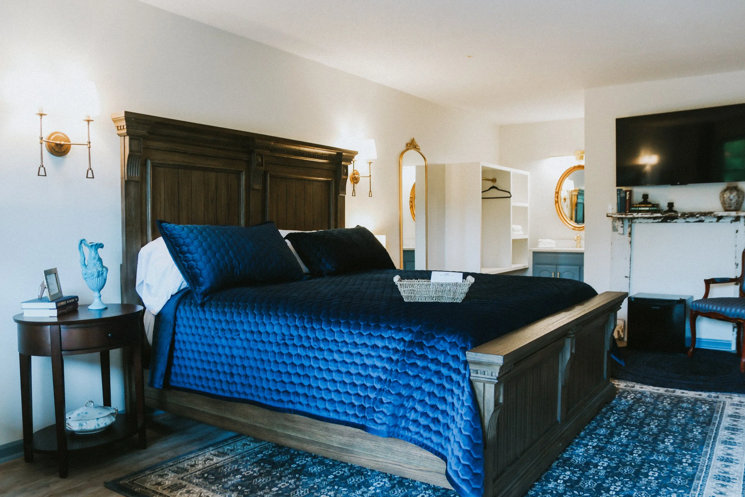 A bedroom with a large wooden bed, navy blue quilt and pillows, side table with books and decorative blue pitcher, wall-mounted lights, white walls, a flat-screen TV, a black and white patterned rug, and an open closet with a gold-framed mirror.