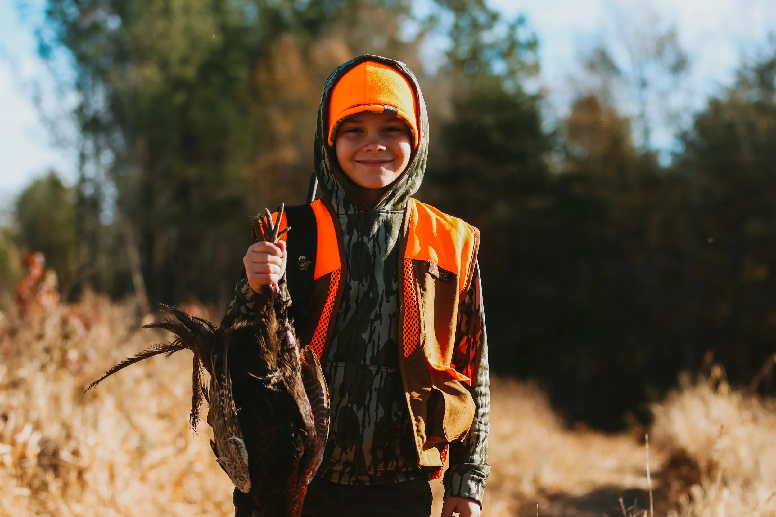 A young boy standing outdoors in a field holding a hunting or fishing shotgun and a recently caught game bird, wearing a camouflage hoodie and a bright orange safety vest and hat, with trees in the background.