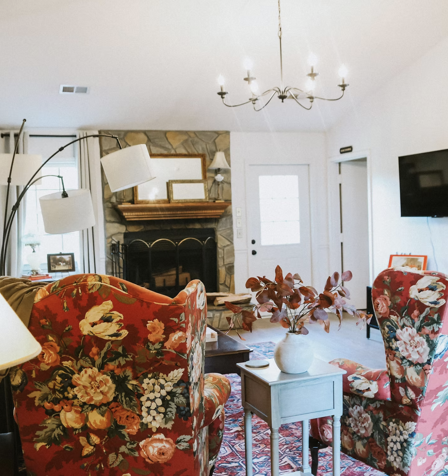 Living room with red floral upholstered armchairs, a fireplace with a stone surround, a chandelier, a door with a window, a wall-mounted TV, and a side table with a vase of red leaves.