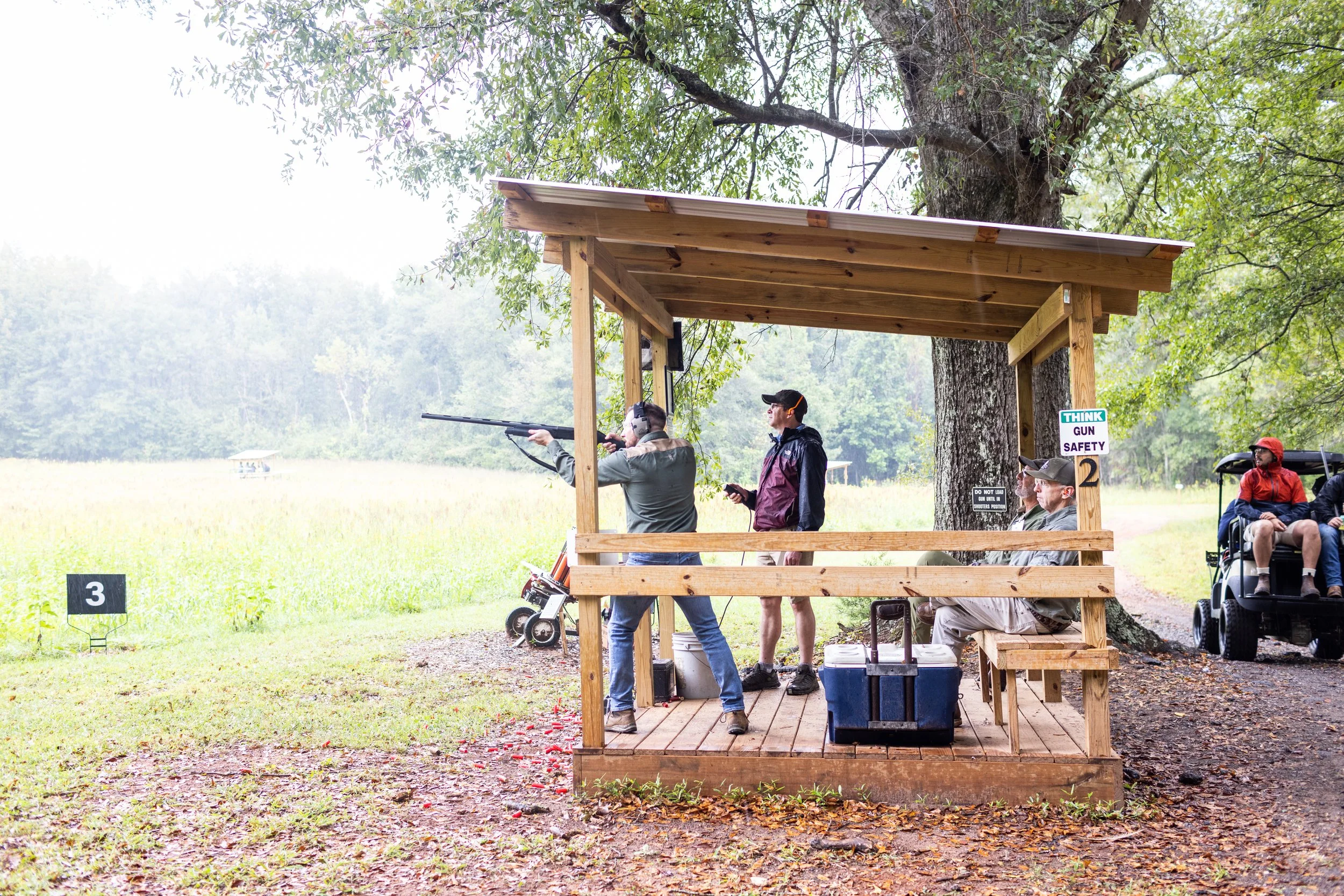 People shooting clay targets at a shooting range under a wooden shelter amid trees.