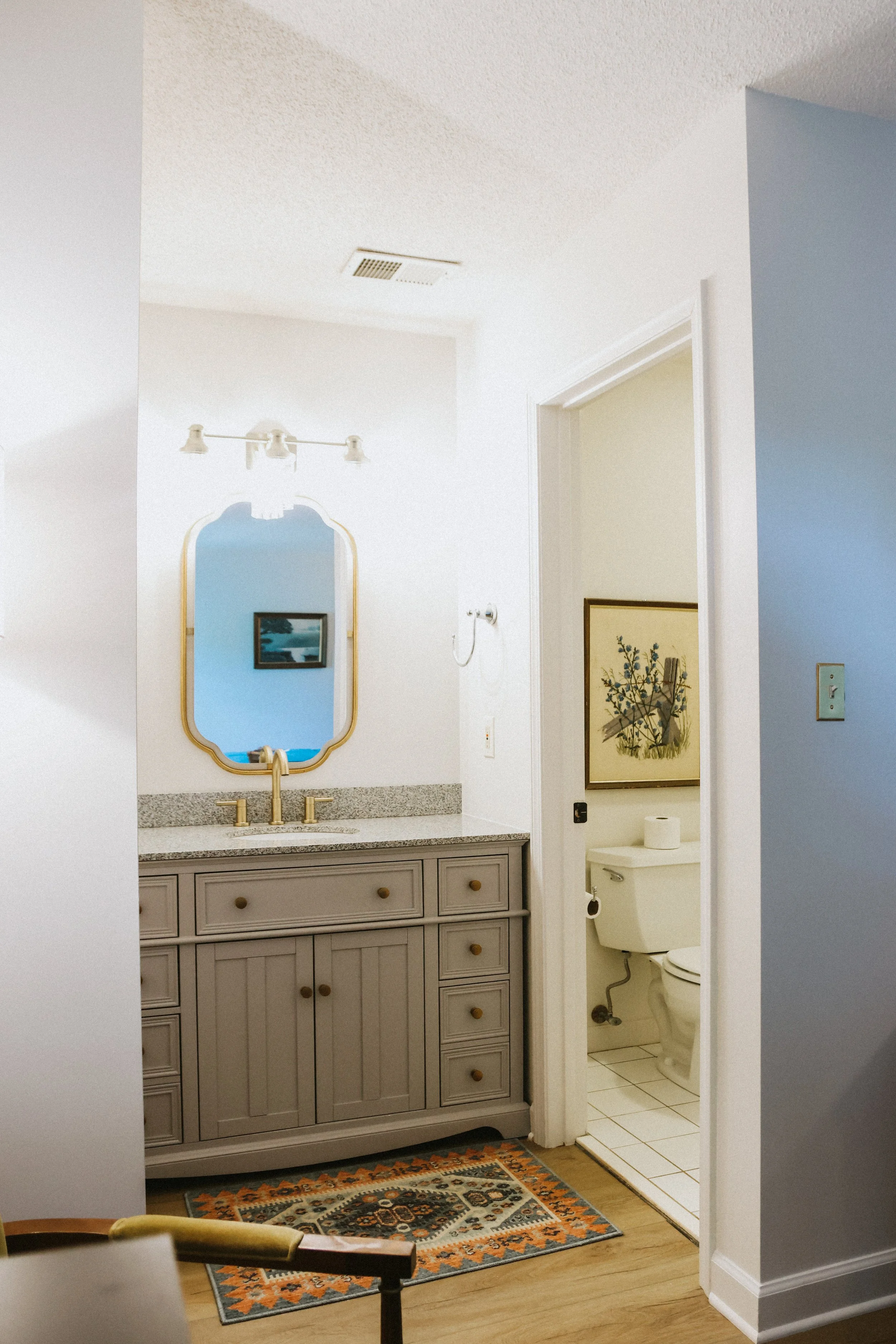 Bathroom with a vanity mirror, sink, beige cabinetry, and a door leading to a toilet area with framed art and a toilet paper roll.
