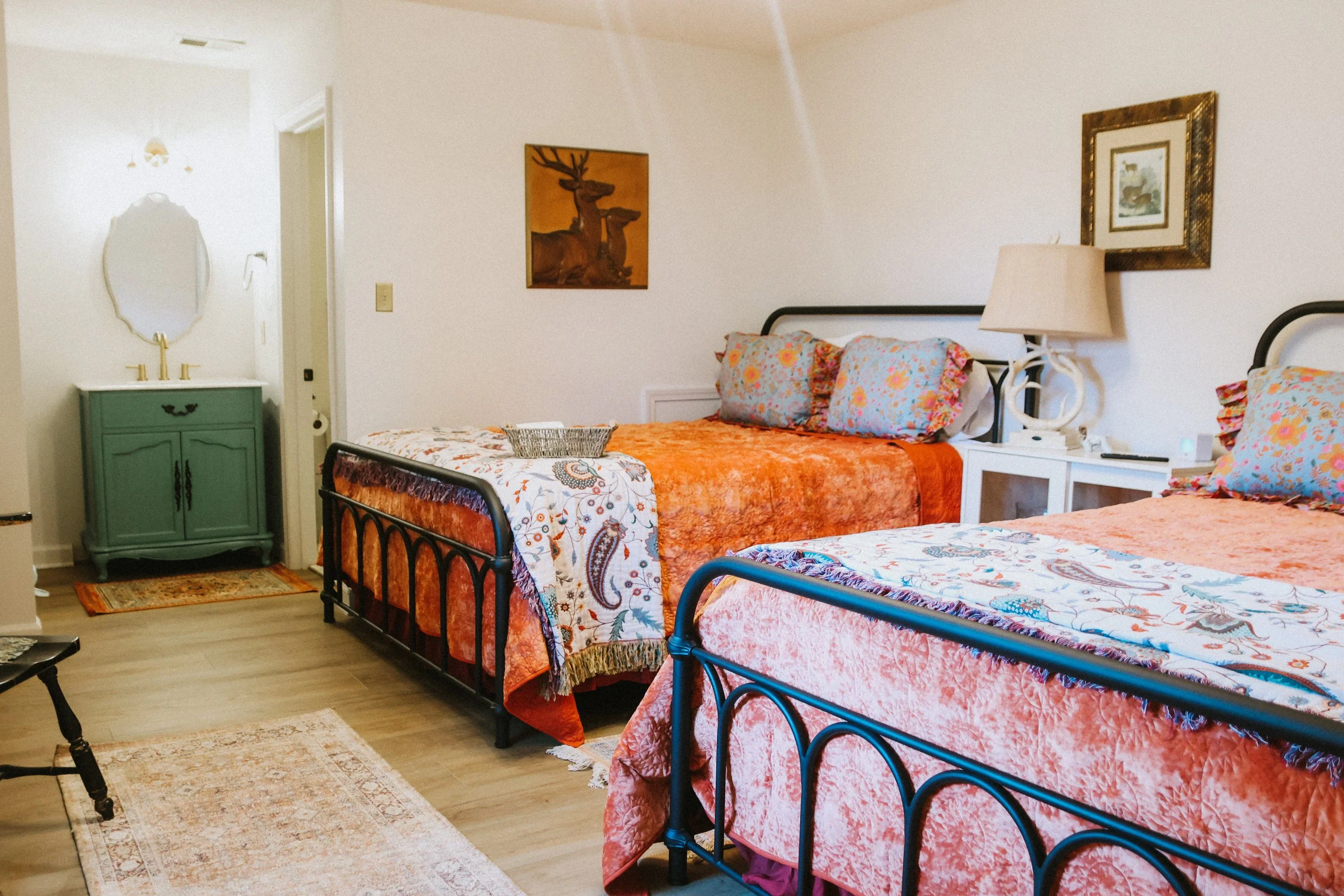 A bedroom with two beds covered in orange and floral bedding, each with floral pillows. There is a white nightstand between the beds with a beige lamp. On the wall above the bed is a framed picture, and another picture of a deer is on the wall to the