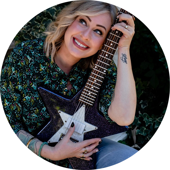 A smiling woman with blonde wavy hair, wearing a colorful patterned blouse, holding an electric guitar with a star shape, and posing outdoors with greenery in the background.