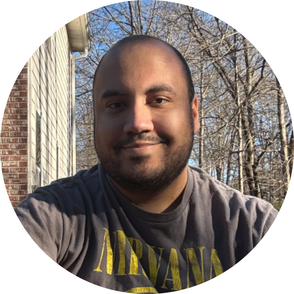 A man with a beard and short hair taking a selfie outdoors on a sunny day, with bare trees and a house in the background.