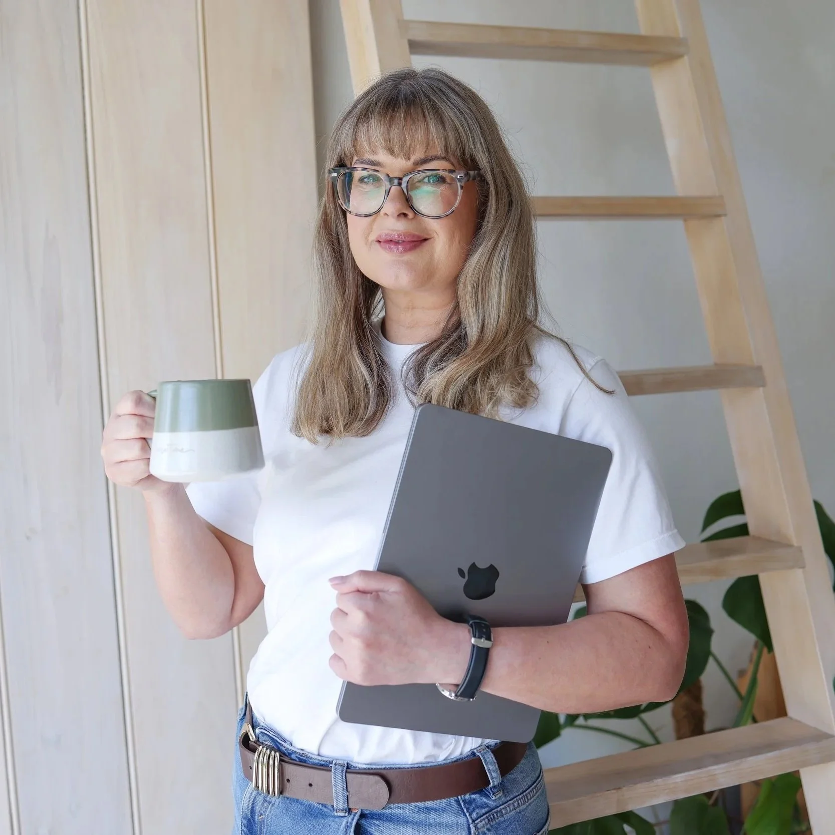 A woman with glasses and long hair holding a laptop and a coffee mug, standing indoors near a wooden ladder and a plant.