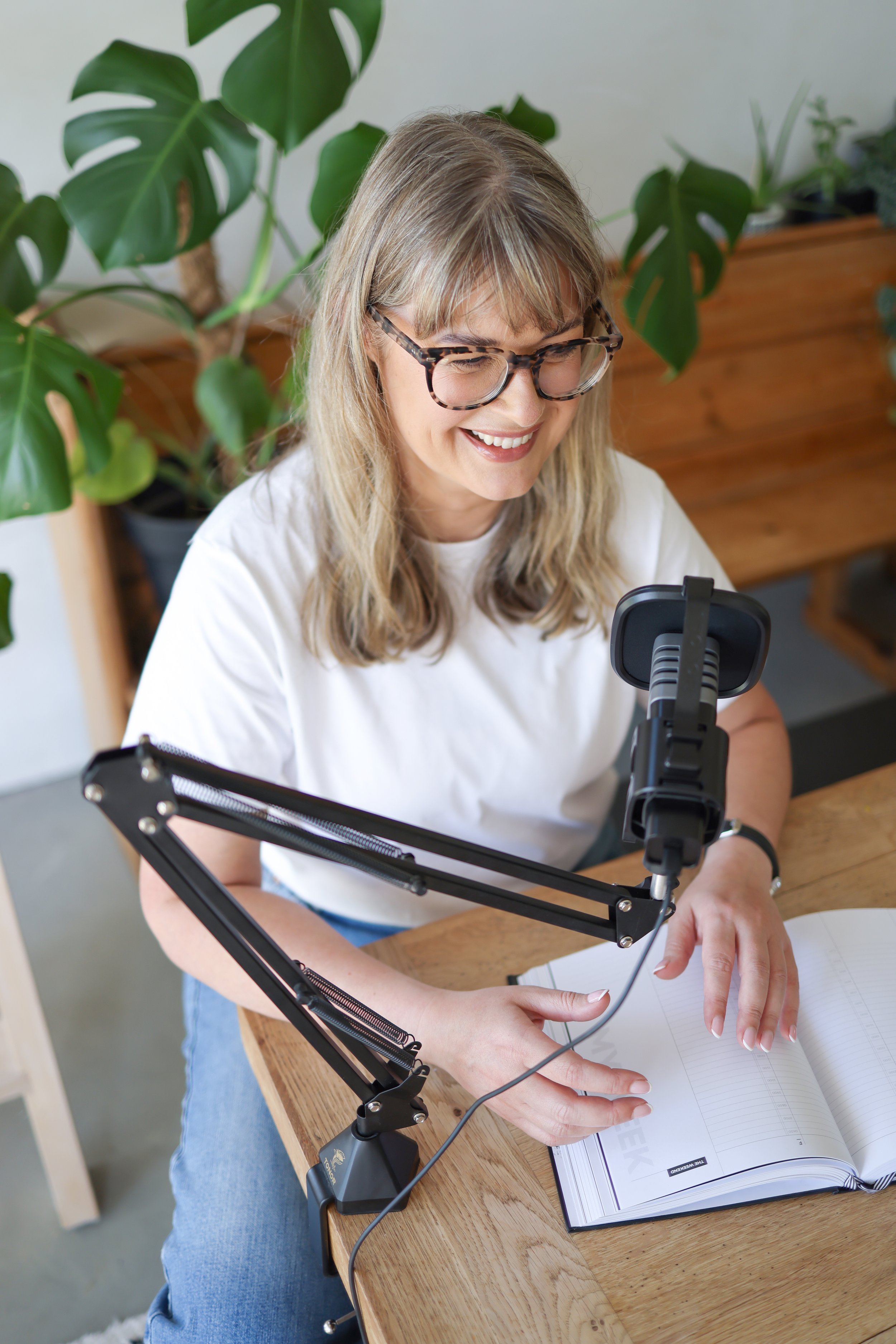 A woman with glasses sitting at a wooden table, smiling while recording herself with a smartphone mounted on a flexible arm, with an open notebook in front of her and a large green plant in the background.