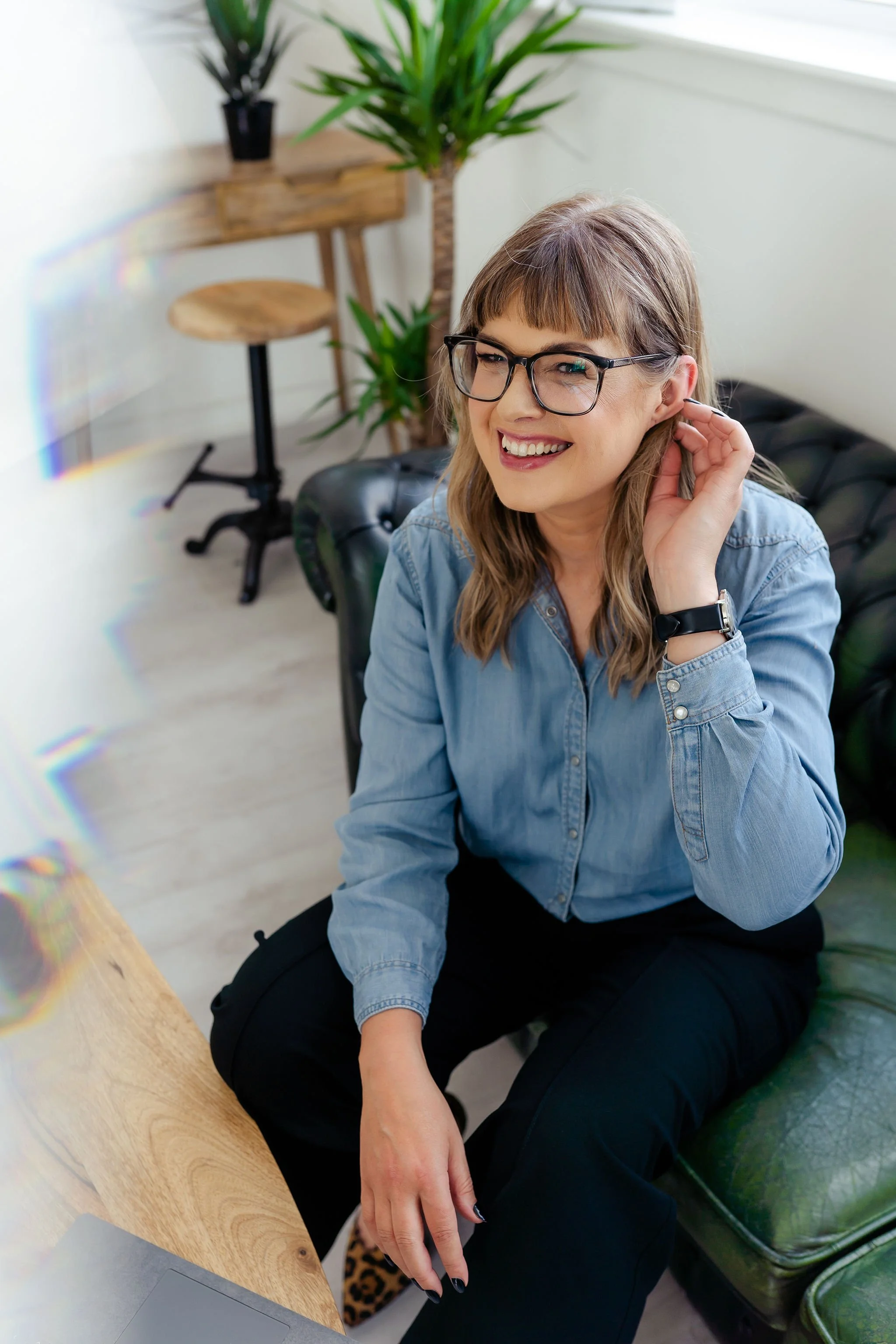 A woman with glasses and long hair, wearing a denim shirt and black pants, sitting on a green sofa in a modern room with plants and a wooden desk.
