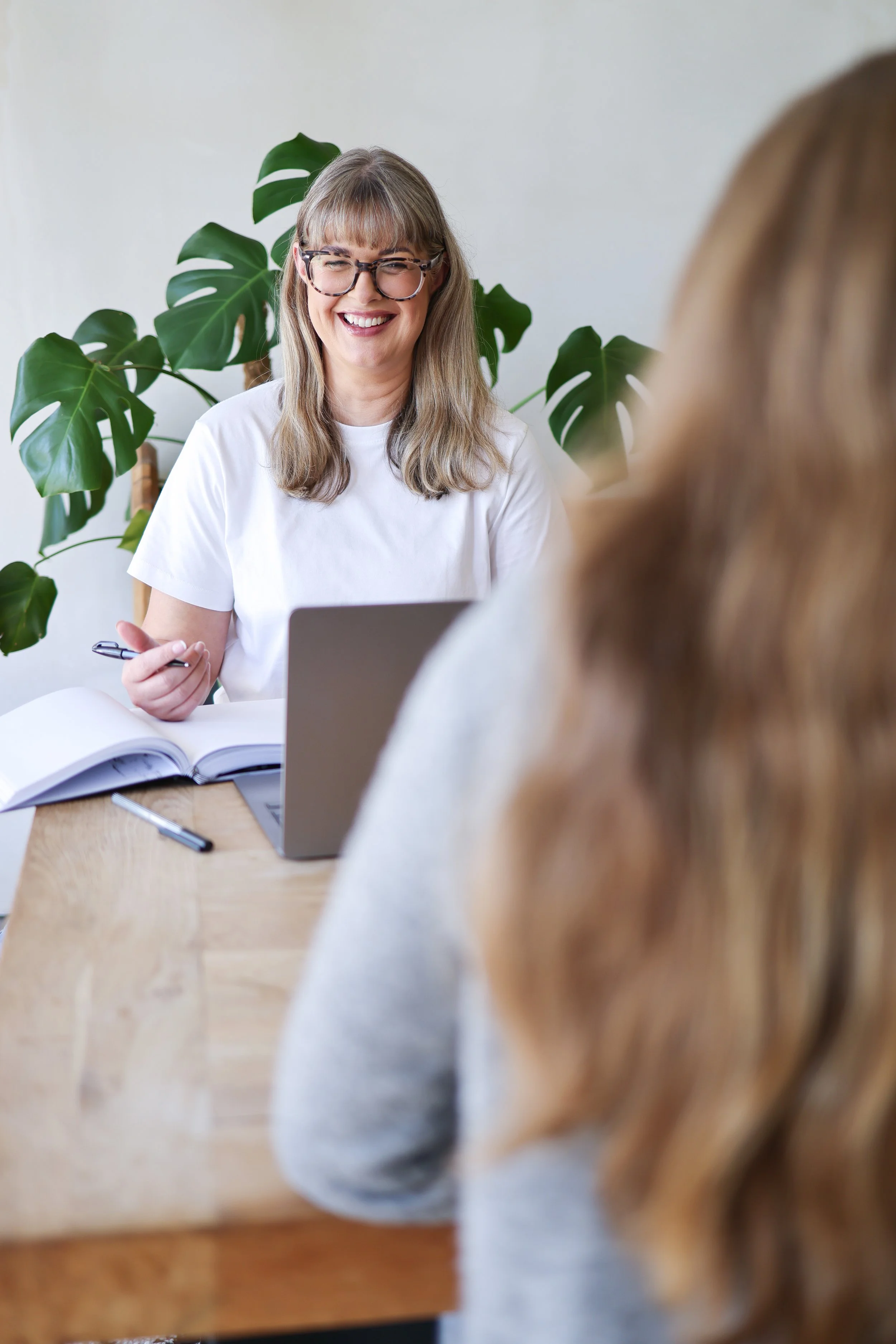 A woman with glasses and long light brown hair is smiling while sitting at a table during a meeting or interview with another woman whose back is to the camera. The woman is wearing a white t-shirt, and there is a laptop, an open notebook, and a pen on the table. Green plants are visible in the background.