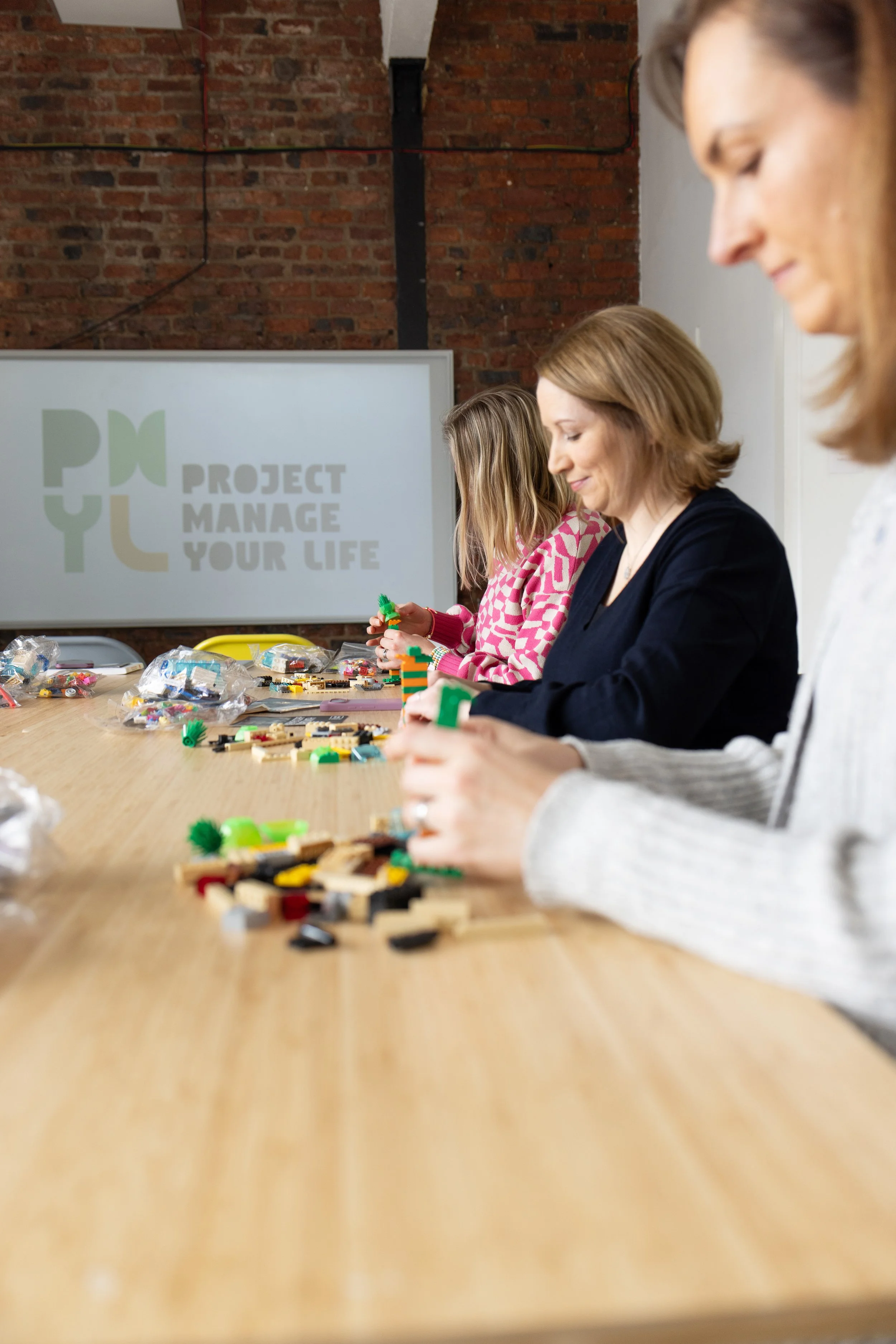 Three women sitting at a large table assembling small building blocks, with a projector screen in the background that reads 'Project Manage Your Life'.