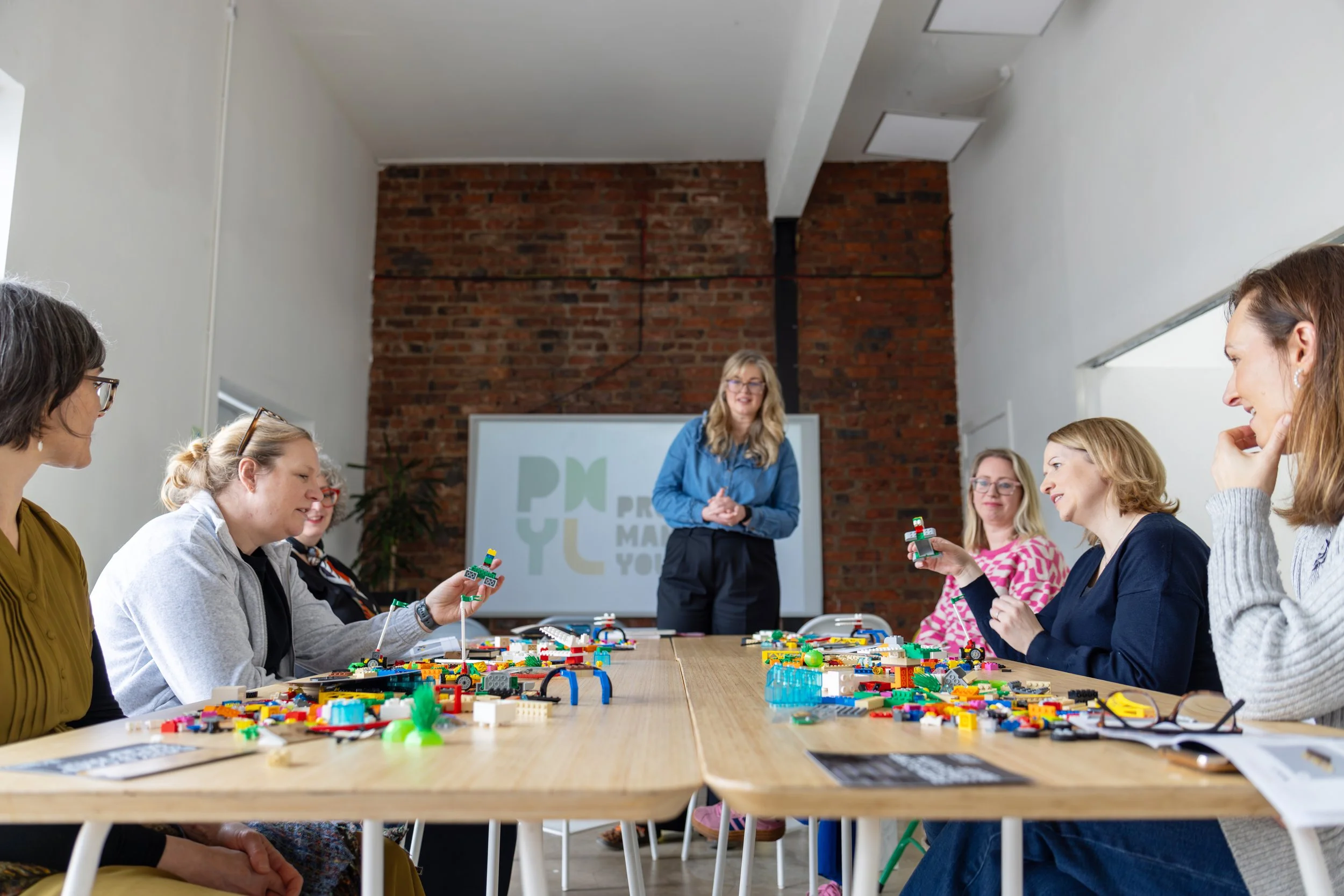 A woman leading a workshop with six women sitting at a long table, building with LEGO bricks. The setting is a room with white walls, a brick accent wall, and a whiteboard in the background.