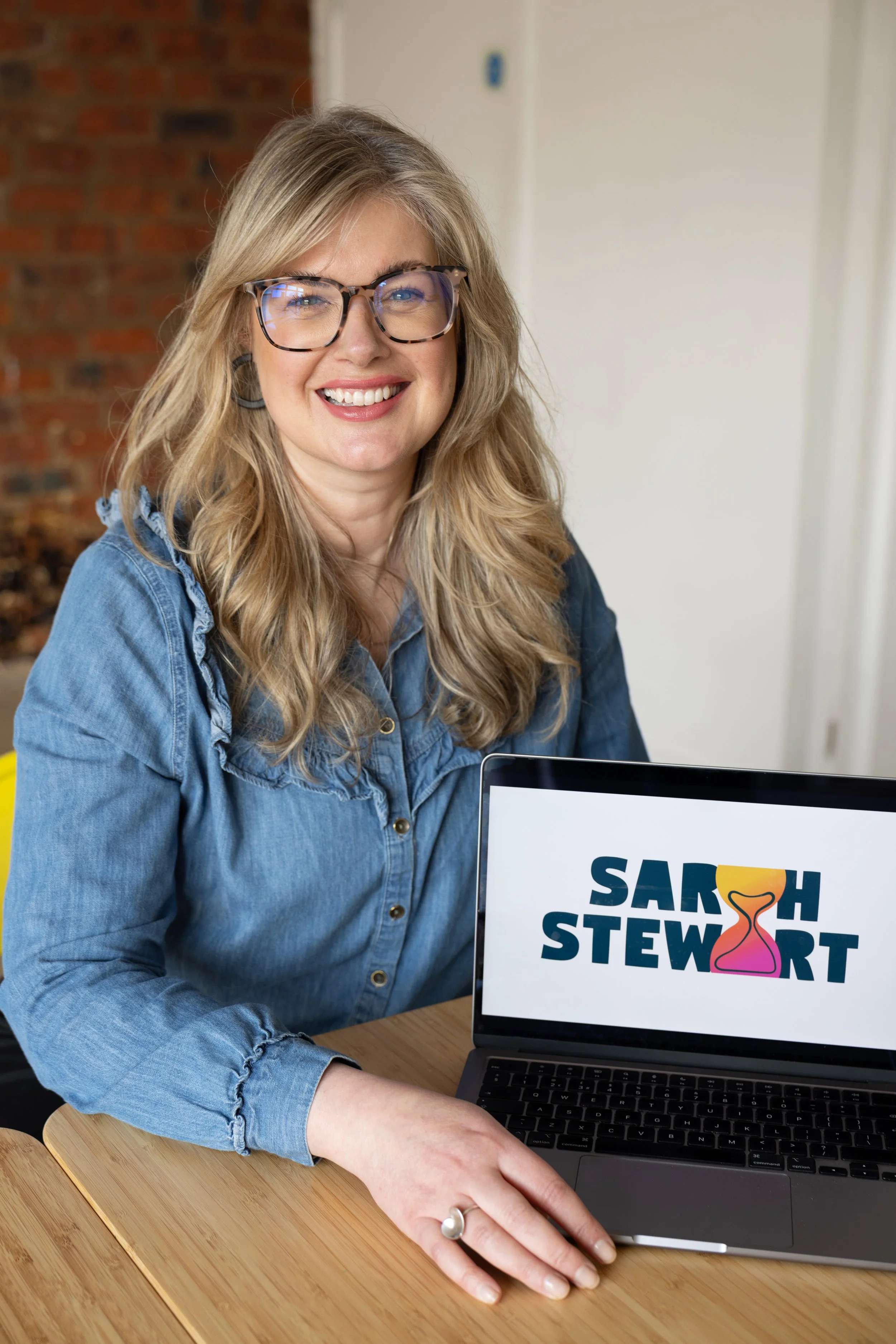 A woman with blonde hair, glasses, and a denim shirt smiling at the camera, sitting at a wooden table with a laptop displaying the name 'Sarah Stewart' with a colorful hourglass logo.