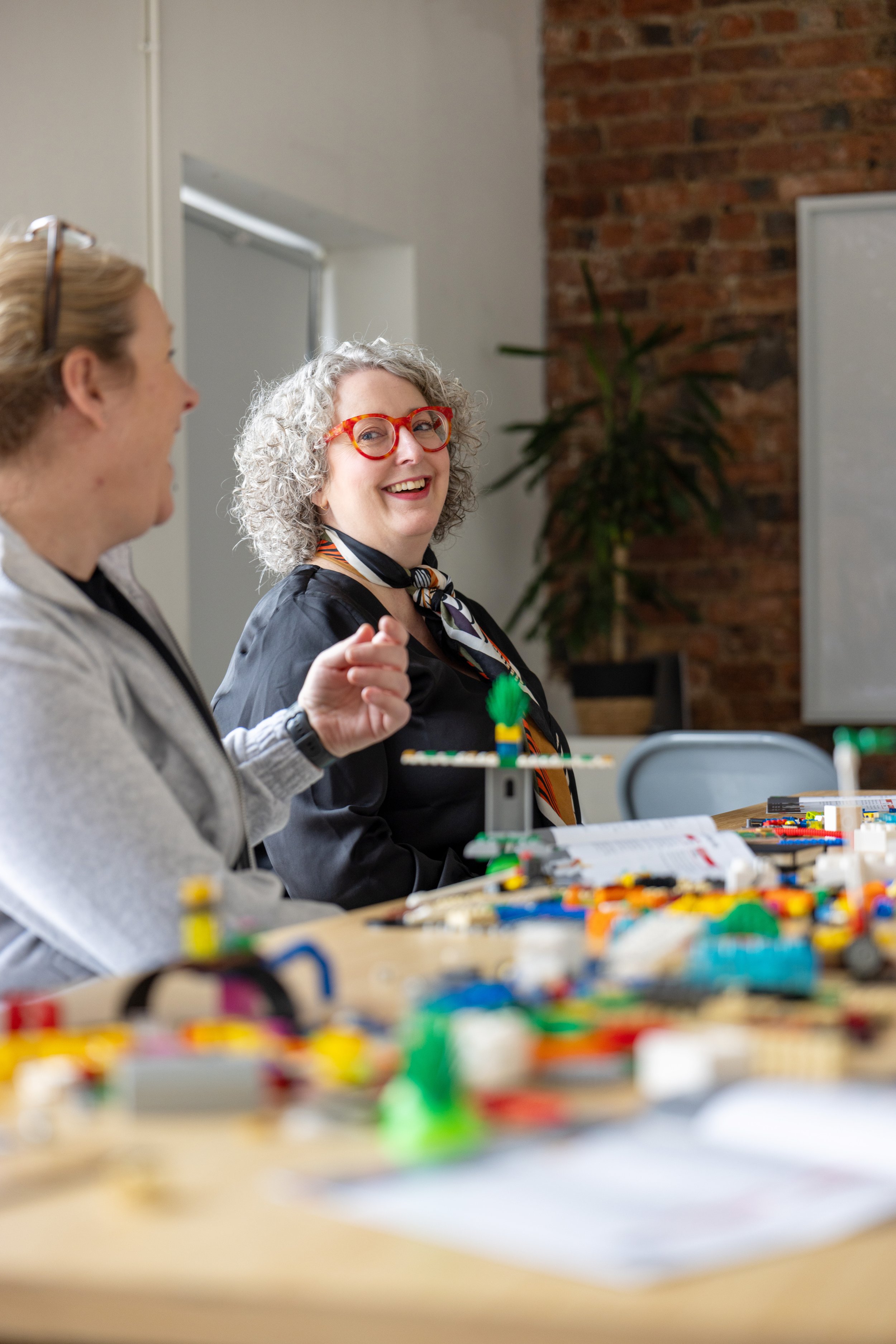 Two women sitting at a table with LEGO bricks, smiling and talking in a casual indoor setting.