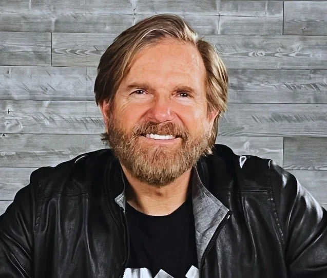A smiling man with a beard and medium-length hair, wearing a black leather jacket over a black t-shirt, sitting against a gray wooden wall.