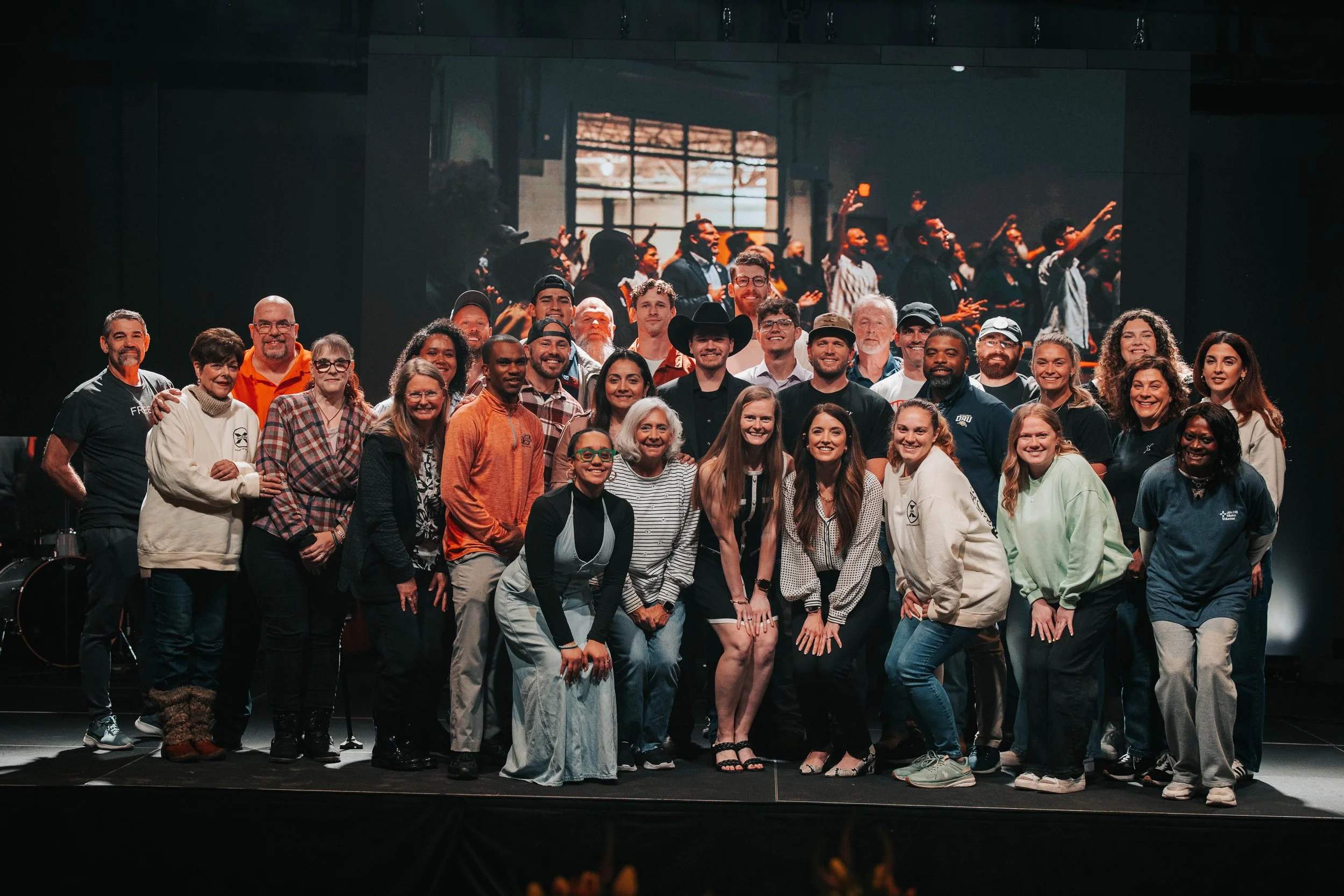 Group of diverse people gathered on stage for a photo, with a dark background and a large screen showing a similar group in an indoor setting.