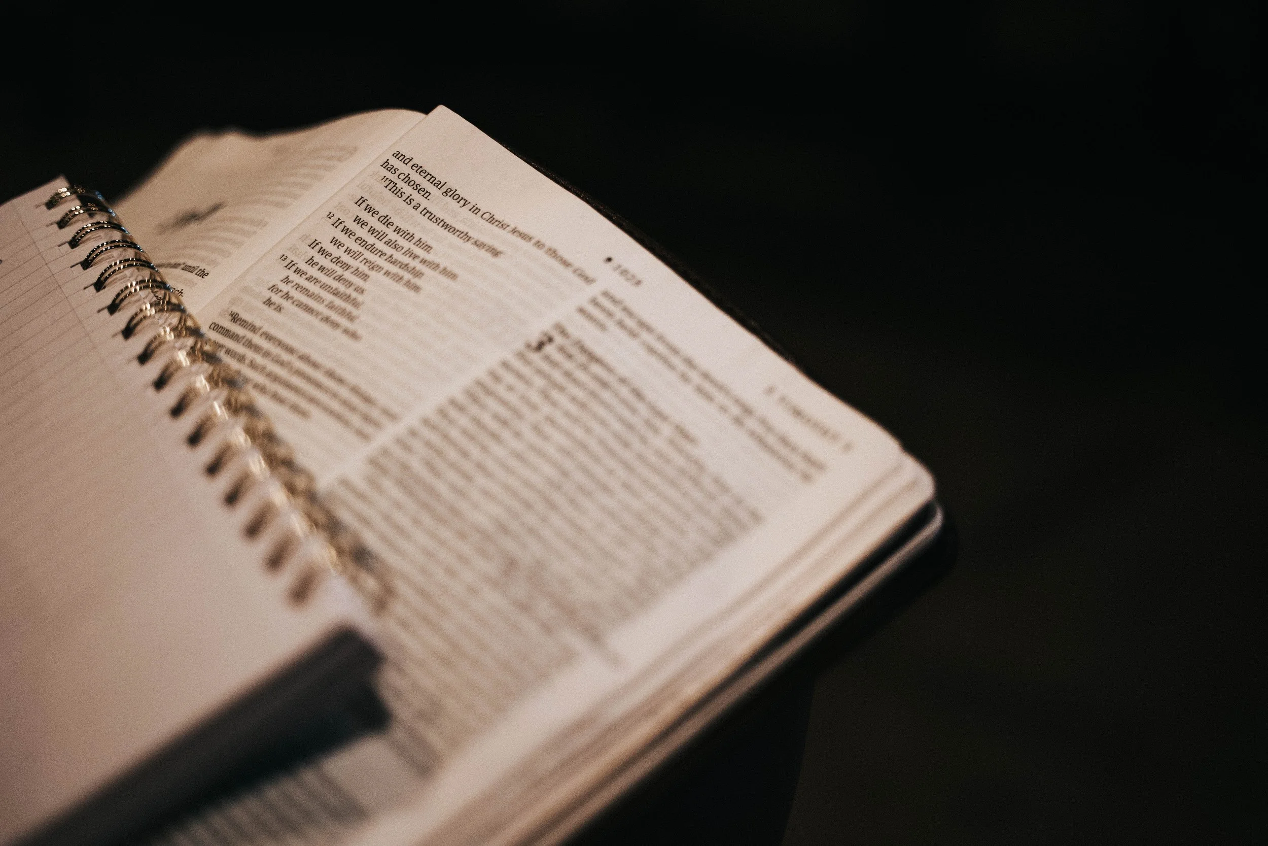 Open spiral-bound book with text on pages, placed on a dark surface, with focus on the left page and text including numbered verses.