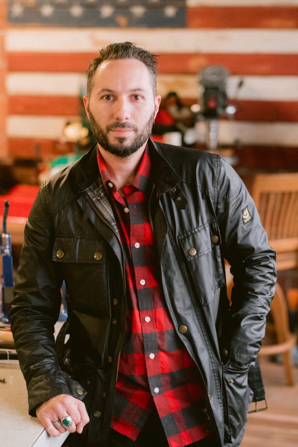 A man with a beard and short hair wearing a black leather jacket over a red and black plaid shirt, standing indoors with an American flag in the background.