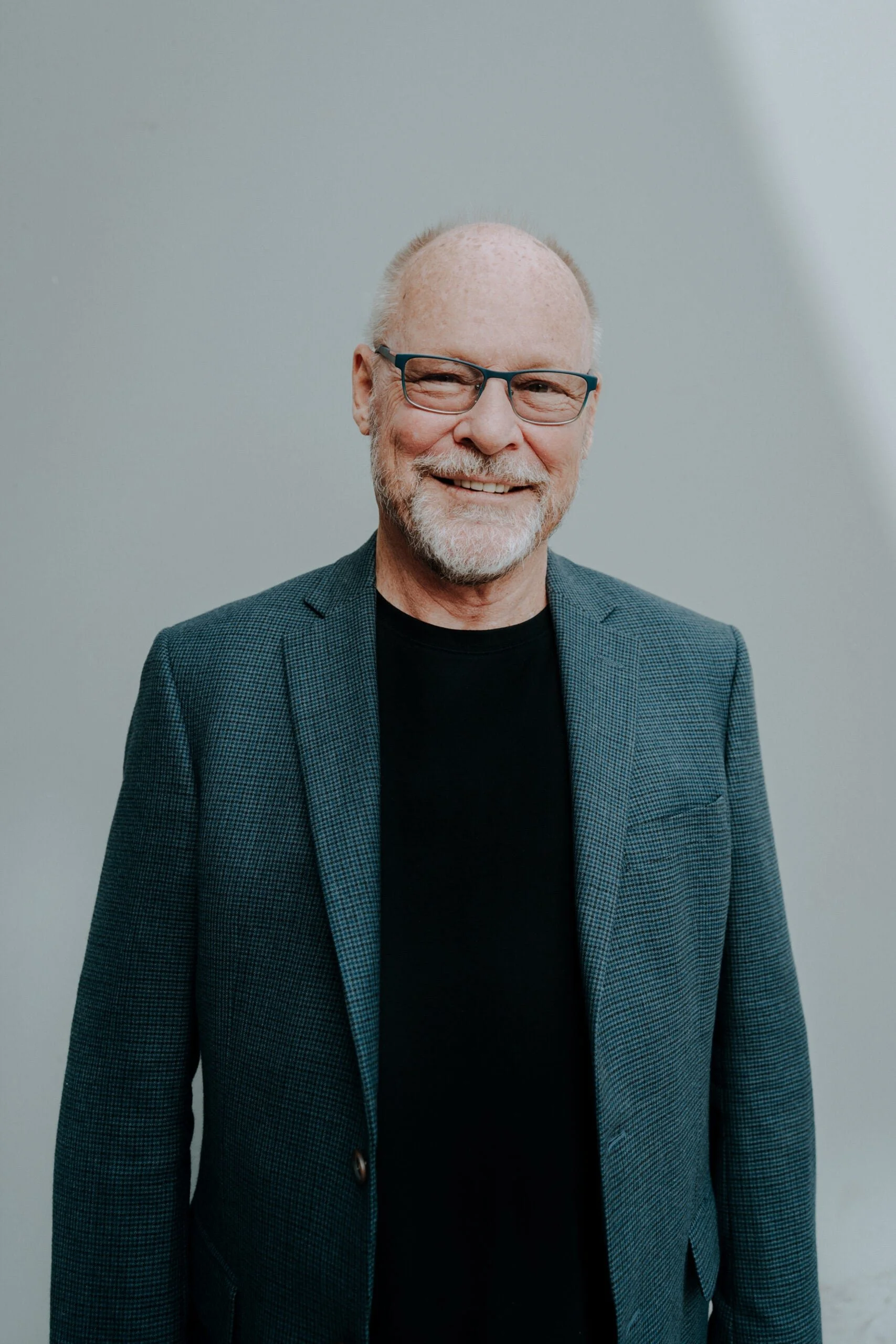 A smiling elderly man with glasses, a beard, and gray hair, wearing a blazer over a black shirt, standing against a plain light gray background.