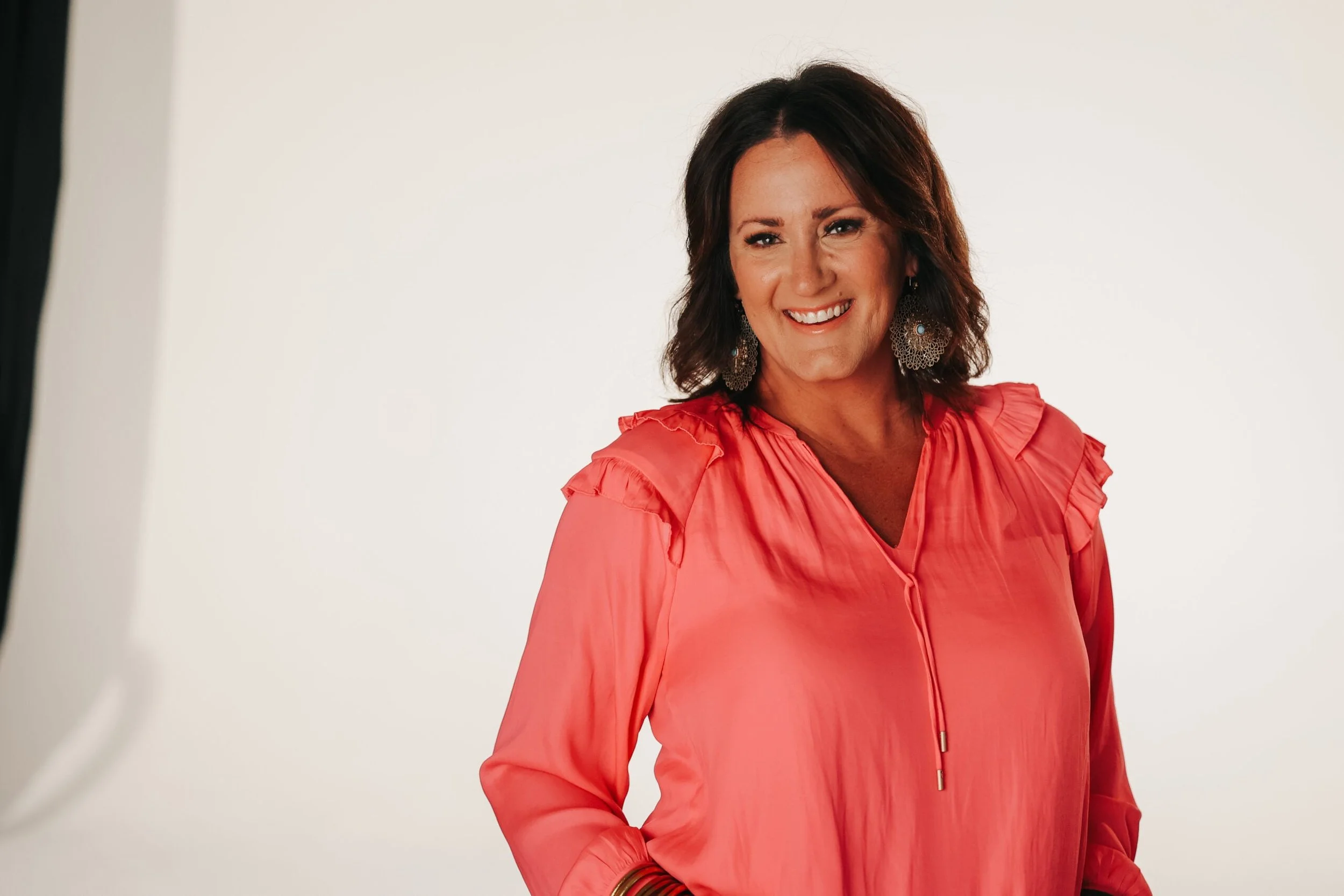 A woman with shoulder-length brown hair wearing a coral pink blouse with ruffled sleeves, large lace-like earrings, and a gold bracelet, smiling against a plain white background.