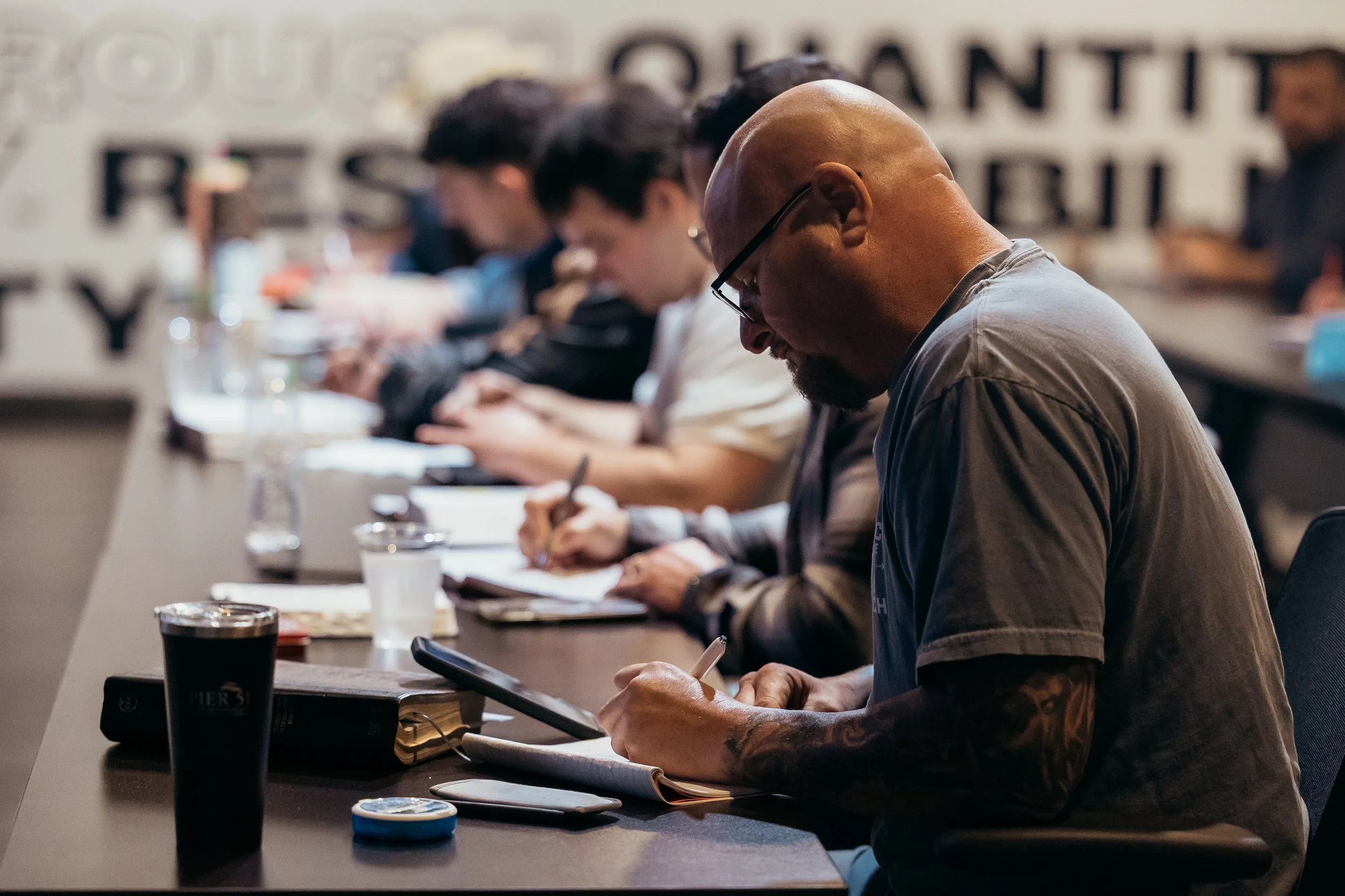 Man with glasses and tattoos wearing a gray shirt sitting at a conference table, writing in a notebook, with other people beside him also engaged in note-taking or writing.