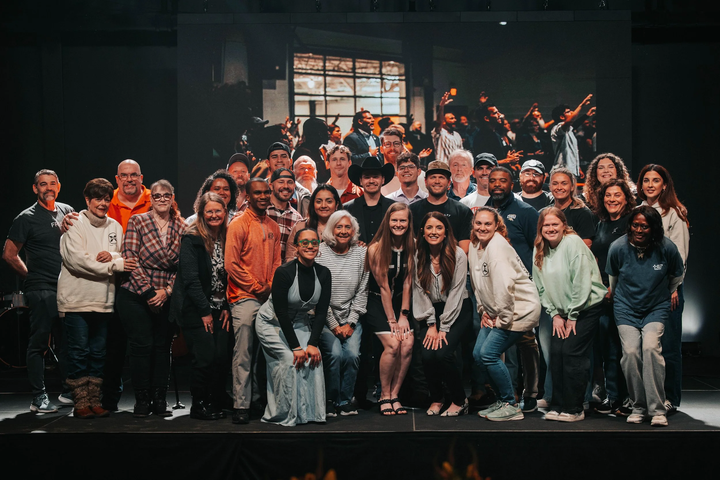 A large group of people posing together on stage, smiling, with a background projection showing others with raised hands in a church or community center setting.
