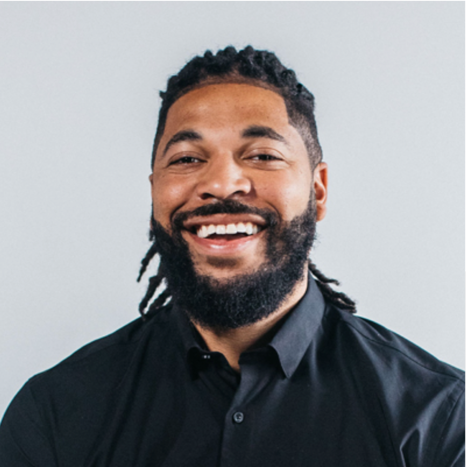 A smiling man with dreadlocks and a beard wearing a black collared shirt against a gray background.