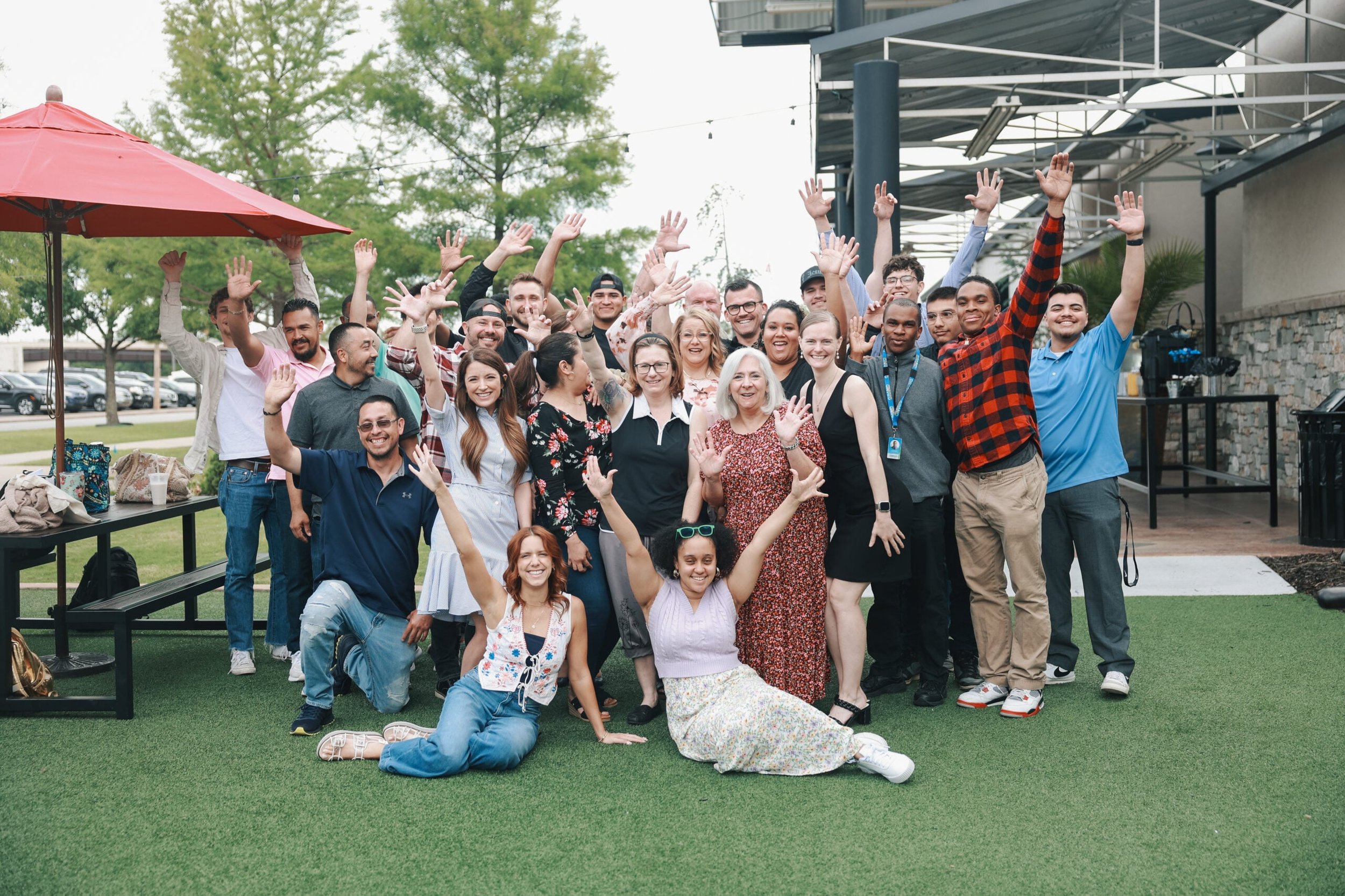 A diverse group of smiling people posing for a photo outdoors under a large red umbrella, with some raising their hands, standing on artificial grass near a building with outdoor seating.
