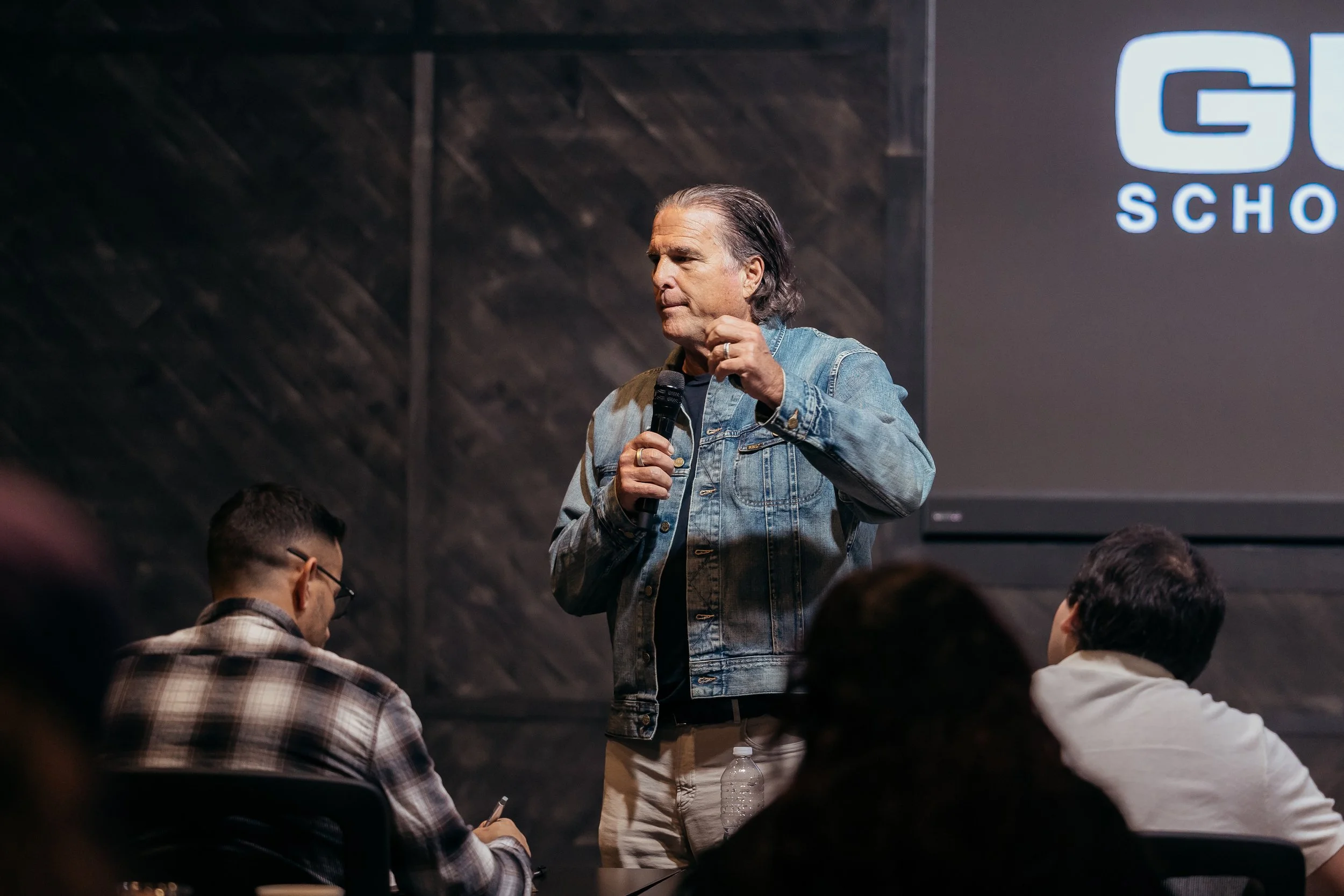 A man with gray hair and a denim jacket speaking into a microphone during a presentation or lecture in front of an audience. There is a large screen behind him with a partial logo and text that says 'G' and 'SCHO'. Audience members are seated, listening attentively.
