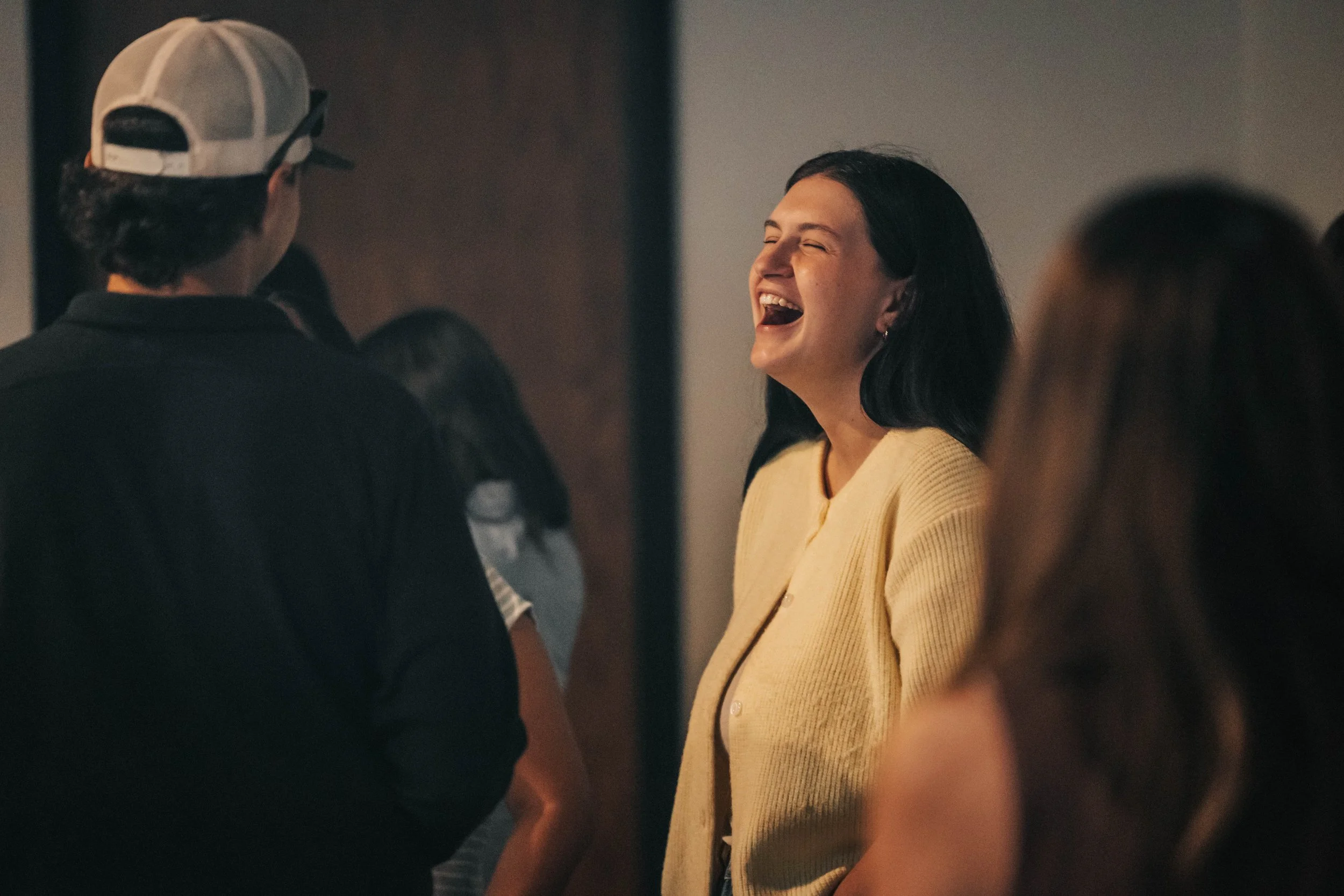 A woman with dark hair, wearing a yellow sweater, is laughing and talking to a person wearing a white baseball cap and dark clothing, in a social setting with other people visible in the background.