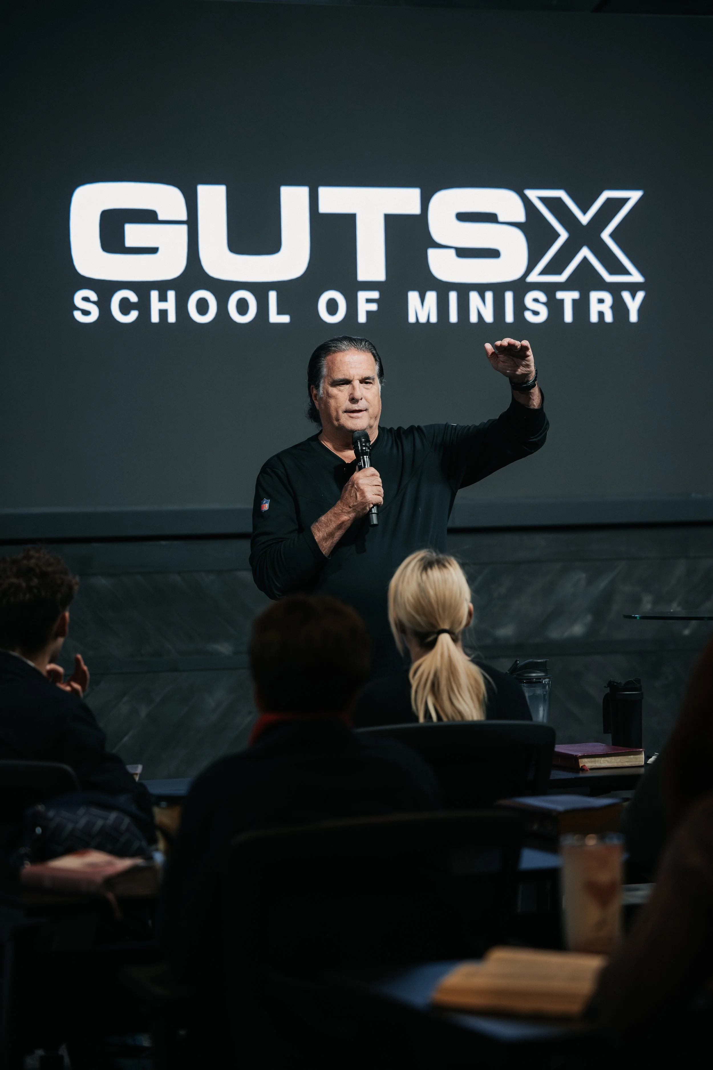 Man speaking into a microphone and gesturing with his hand during a presentation at GUTS X School of Ministry, in front of a large screen displaying the event name.