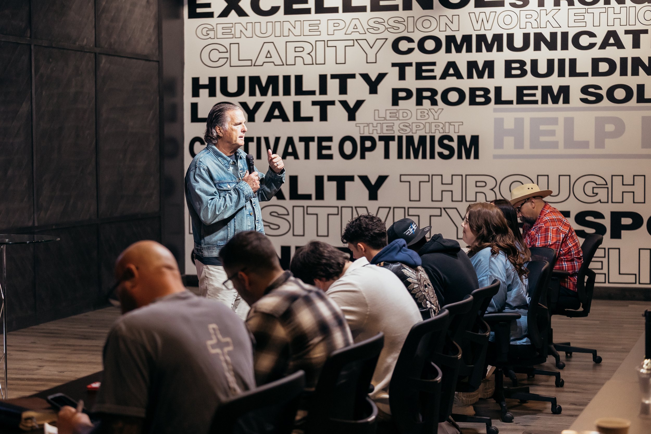 A man in a denim jacket speaking into a microphone in front of a large white wall with motivational words in black and white. Several seated people are listening, some taking notes, in a room with dark wooden wall panels.