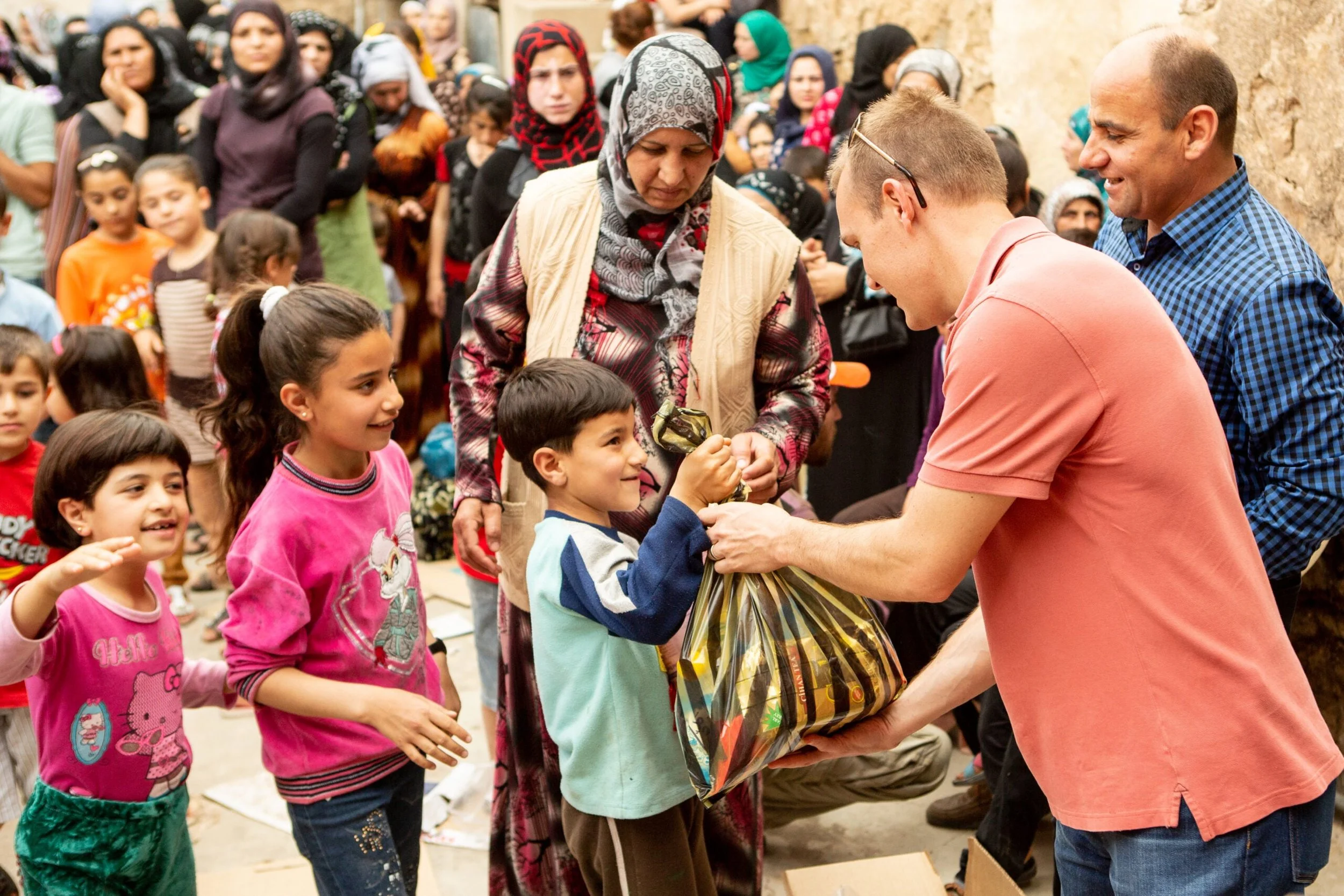 Group of children and adults in a line, receiving gifts from a man in a pink shirt. The scene takes place outdoors, likely in a Middle Eastern setting, with a stone wall in the background. The children are smiling and appear happy.