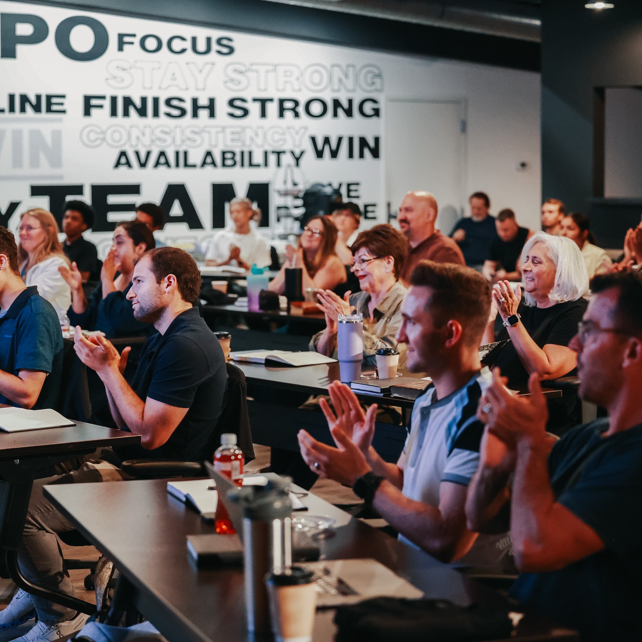 Group of people sitting at tables in a conference room, listening and clapping, with a large motivational words poster on the wall behind them.