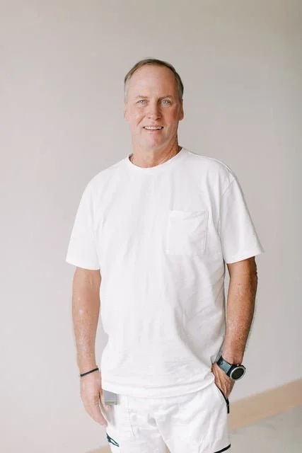 A middle-aged man with short gray hair, wearing a white T-shirt and white pants, standing against a plain light-colored background.