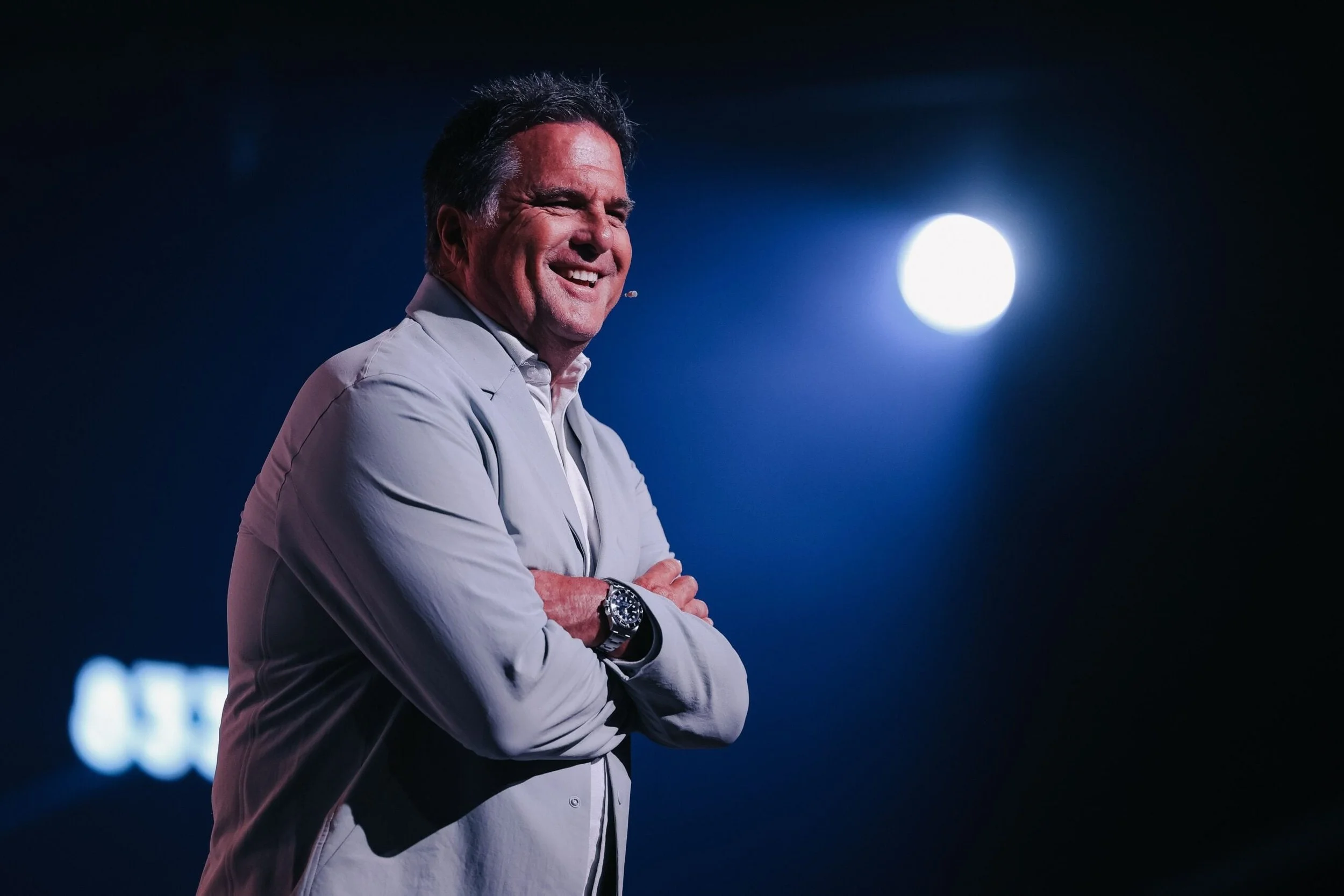 A middle-aged man with dark hair, smiling with his arms crossed, wearing a light grey jacket and silver watch, standing against a dark background with a bright full moon visible behind him.