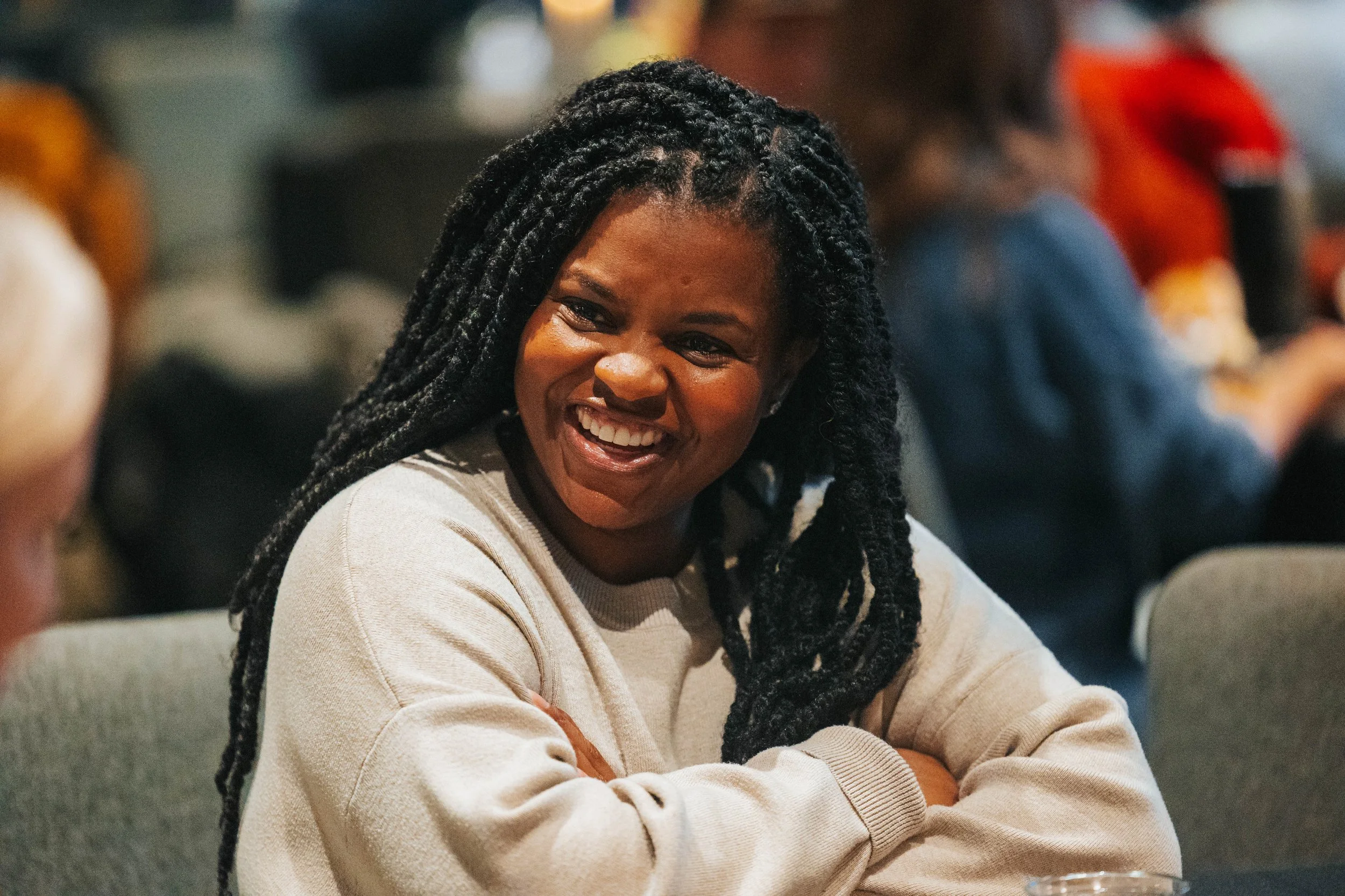 A woman with long braided hair smiling and sitting with her arms crossed in a social setting with blurred people in the background.